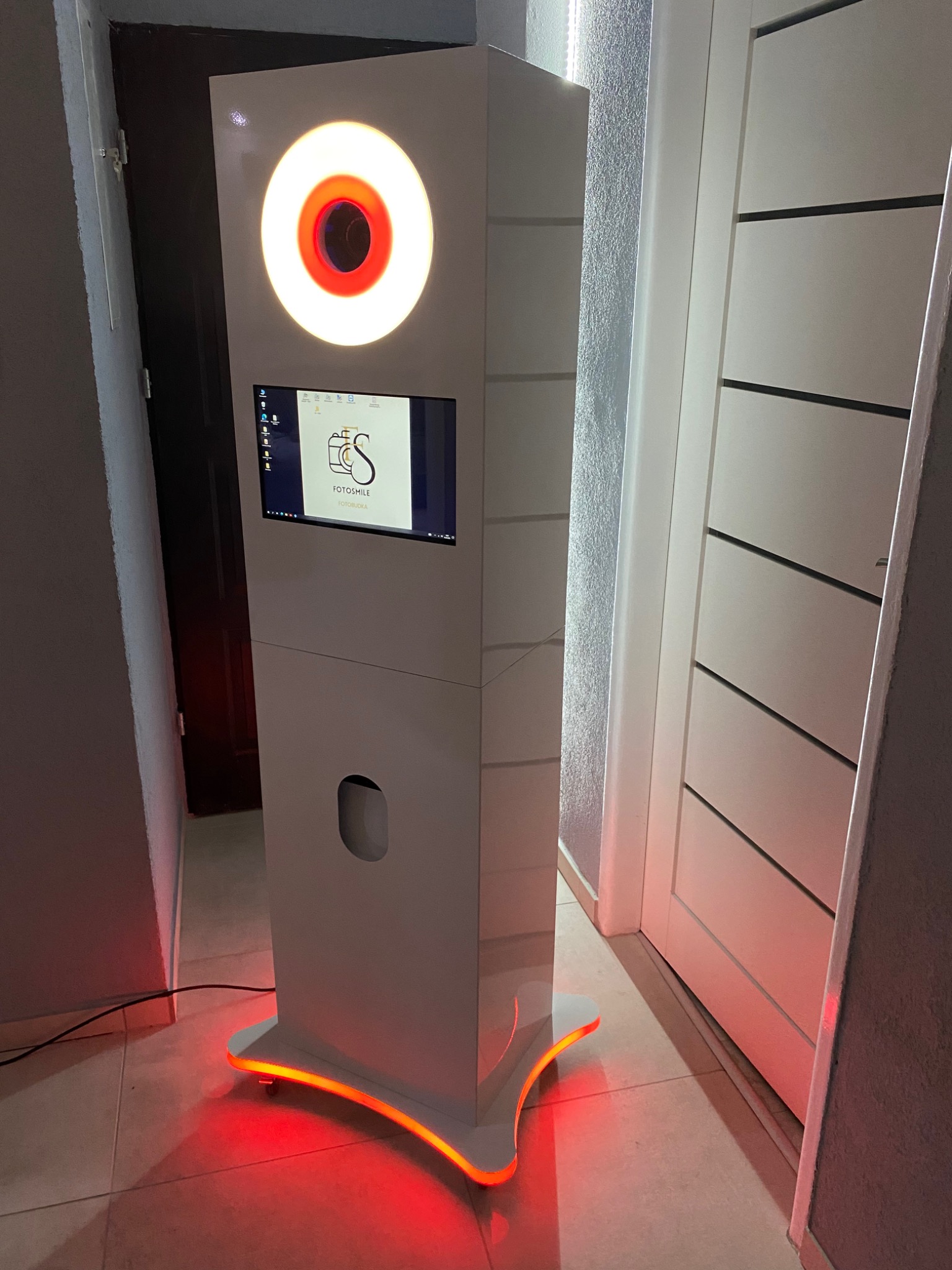 Biała fotobudka z okrągłym, podświetlanym panelem i ekranem dotykowym prezentującym logo firmy Fotosmile, dodatkowo podświetlona od dołu na czerwono.