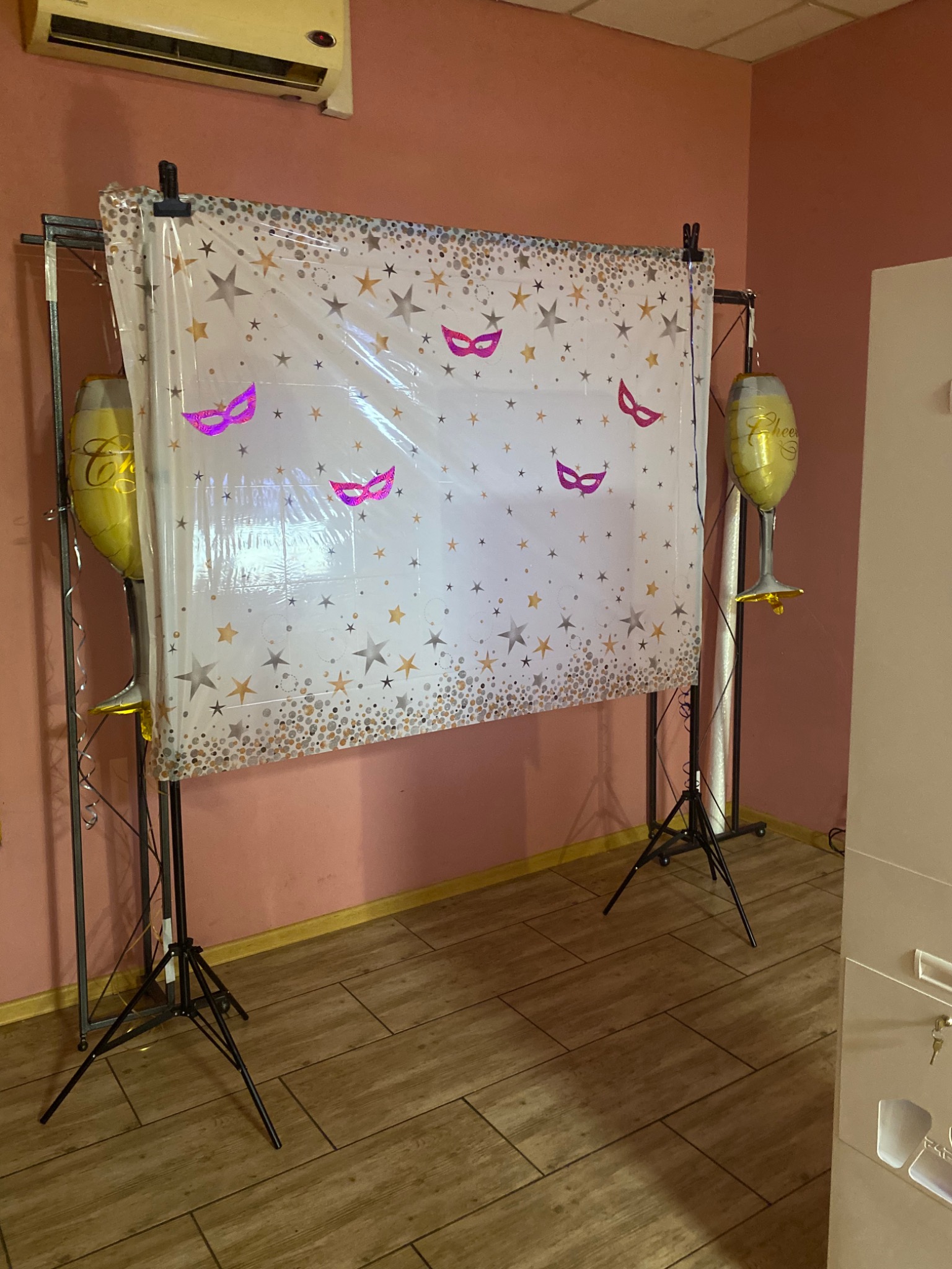 Tło fotograficzne z motywem gwiazd i masek karnawałowych, ustawione na czarnym statywie, z balonami w kształcie kieliszków po bokach, na tle różowej ściany.