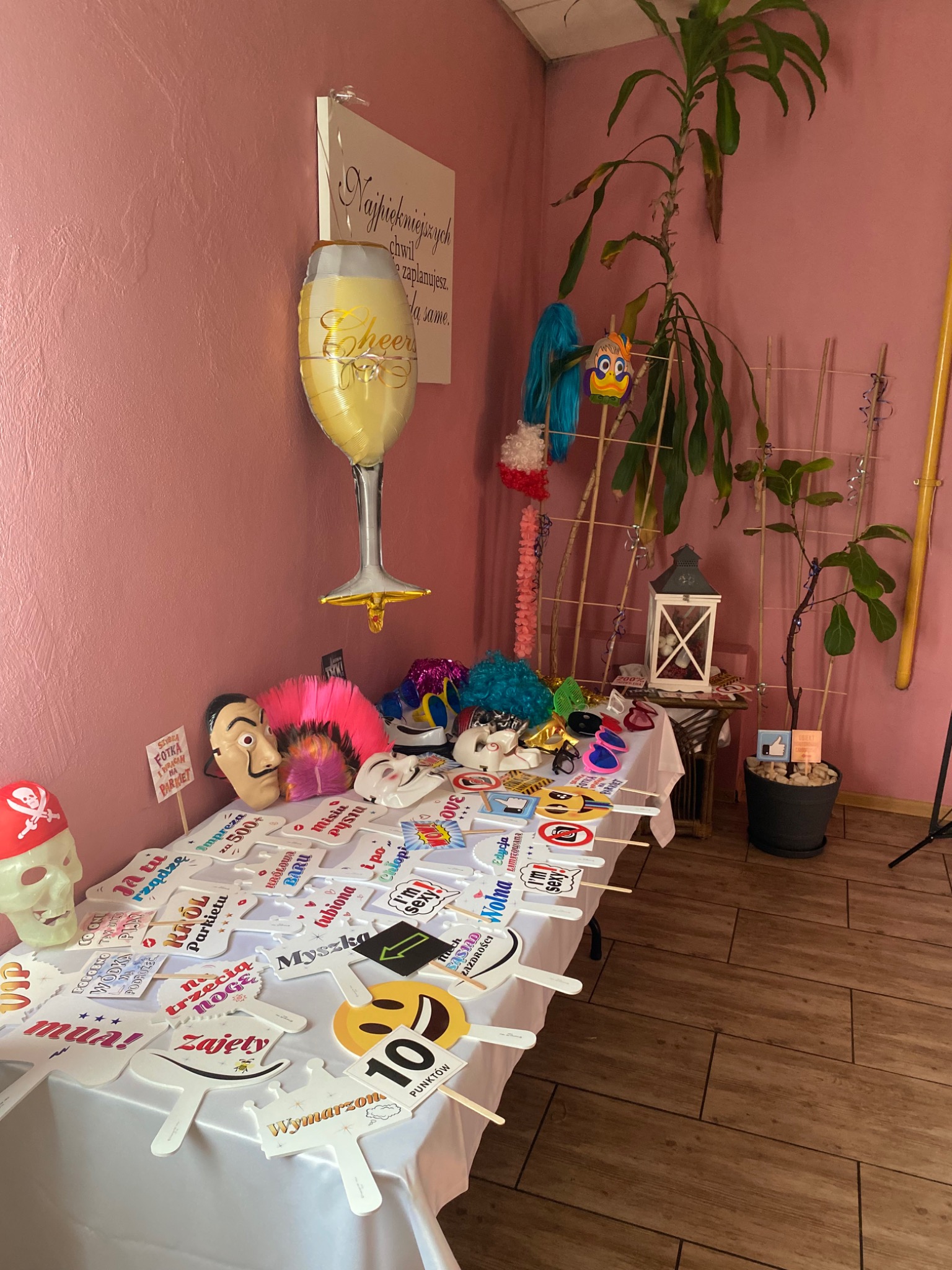 Stół z rekwizytami do fotobudki: maski, peruki, okulary, tabliczki z napisami i balon w kształcie kieliszka na tle różowej ściany i roślin doniczkowych.