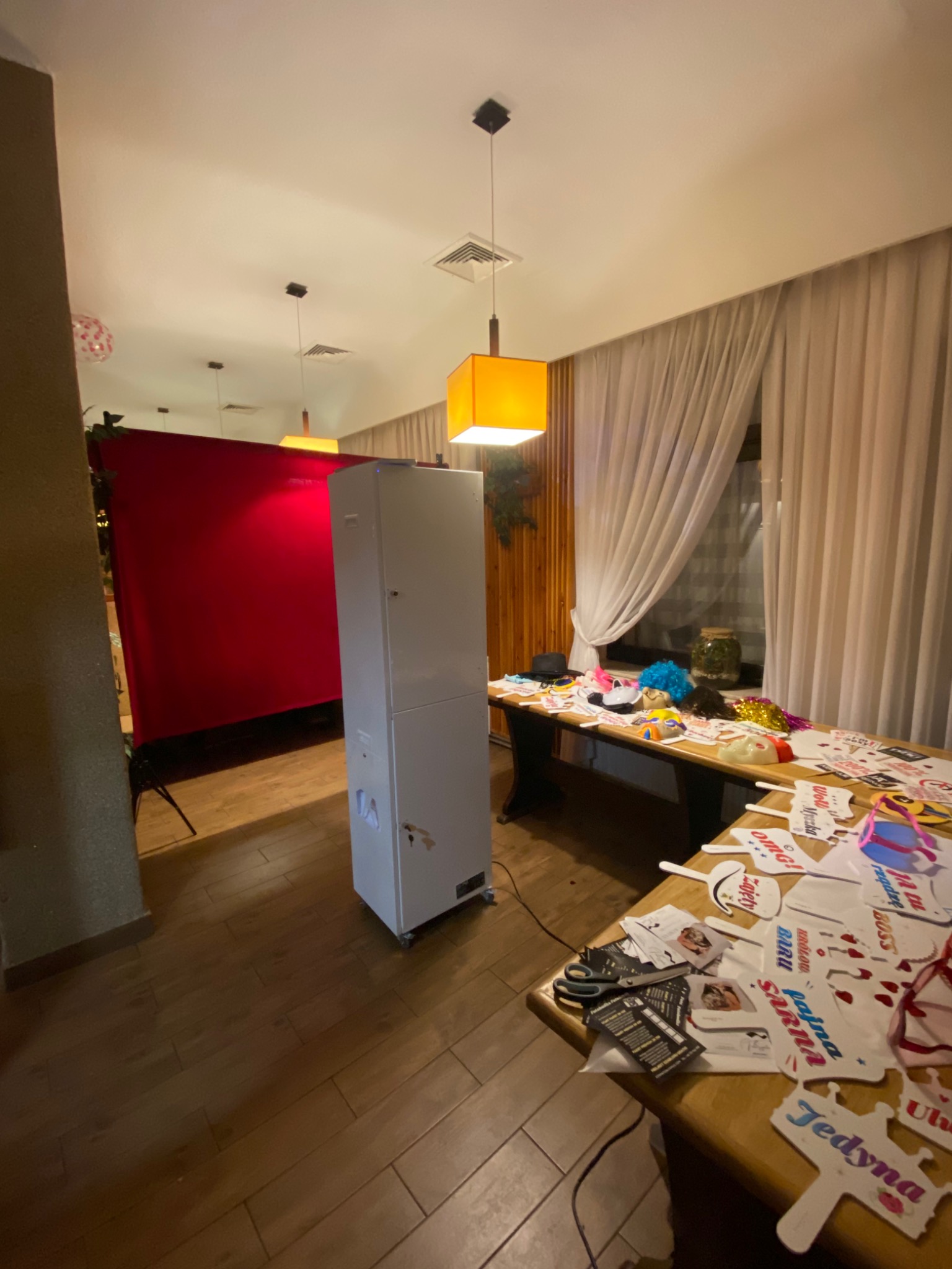 Przygotowania do sesji zdjęciowej: fotobudka z czerwonym tłem, stół z rekwizytami, maskami i napisami na patykach, oświetlona ciepłym światłem lamp.