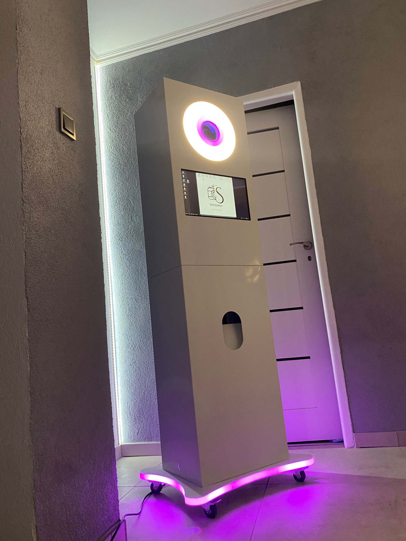 Biała fotobudka z podświetleniem LED na kółkach, ekranem dotykowym i okrągłym oświetleniem, ustawiona w rogu pomieszczenia z szarą ścianą i białą, zamkniętą drzwiami.