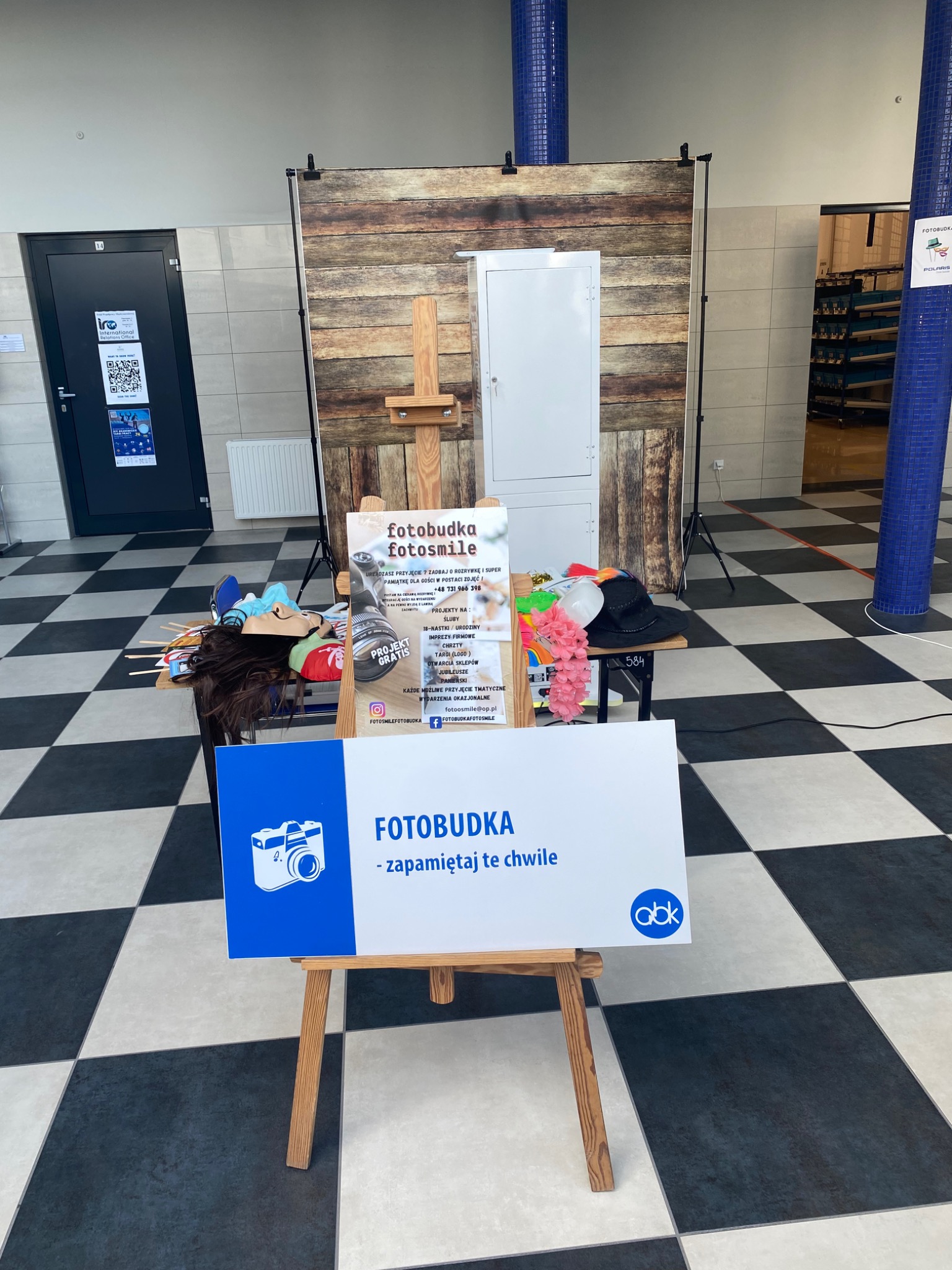 Stanowisko fotobudki z rekwizytami, tłem imitującym drewno i tablicą reklamową na sztaludze, ustawione w przestronnym wnętrzu z szachownicową podłogą.