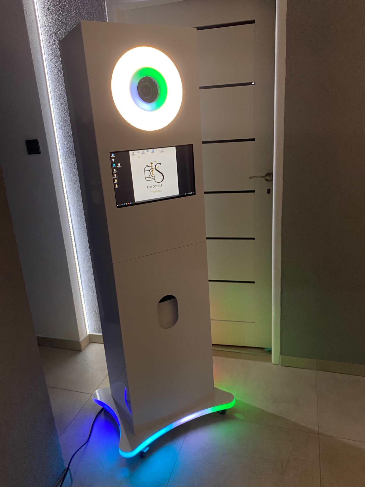 Biała fotobudka z podświetleniem LED w kolorach niebieskim i zielonym, widoczny ekran dotykowy z logo firmy Fotomile oraz oświetlenie pierścieniowe wokół obiektywu aparatu.