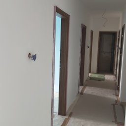 pro-finish Katarzyna Rączka - Korytarz w trakcie malowania, ściany w jasnym odcieniu, widoczne obramowania drzwi z brązowego drewna, zabezpieczona podłoga taśmą malarską, wystające kable ze ściany.