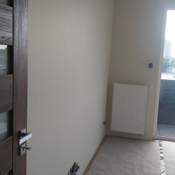pro-finish Katarzyna Rączka - Świeżo pomalowane jasne ściany w pokoju z zakrytą podłogą, widoczne gniazdka elektryczne zabezpieczone taśmą oraz fragment drzwi z ciemnego drewna po lewej stronie.