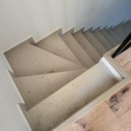 pro-finish Katarzyna Rączka - Widok z góry na wykończone schody z szarych płytek, z widocznymi śladami po remoncie, zabezpieczone białą listwą przypodłogową, z fragmentem drewnianej podłogi i buta w rogu.