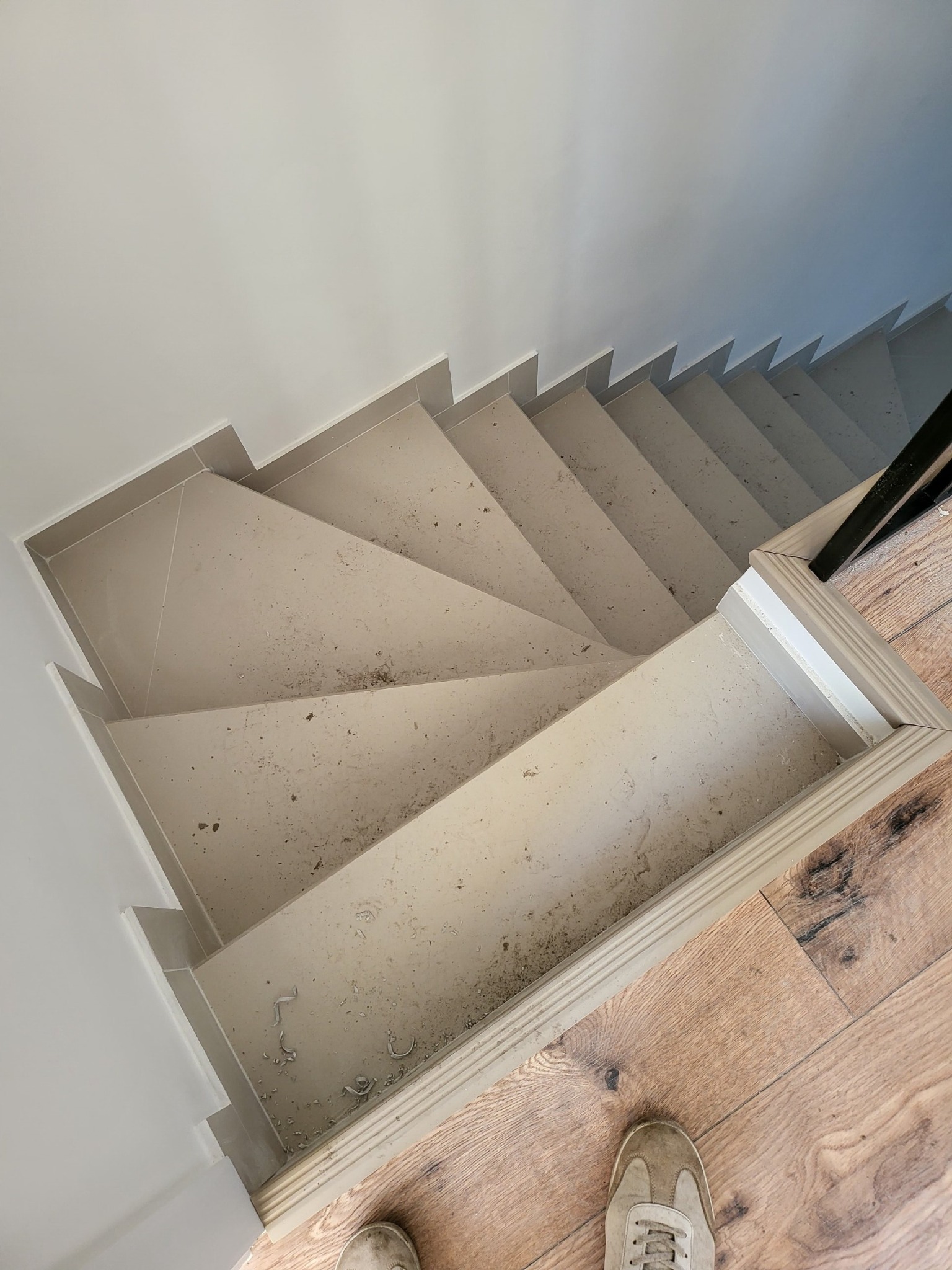 Widok z góry na wykończone schody z szarych płytek, z widocznymi śladami po remoncie, zabezpieczone białą listwą przypodłogową, z fragmentem drewnianej podłogi i buta w rogu.