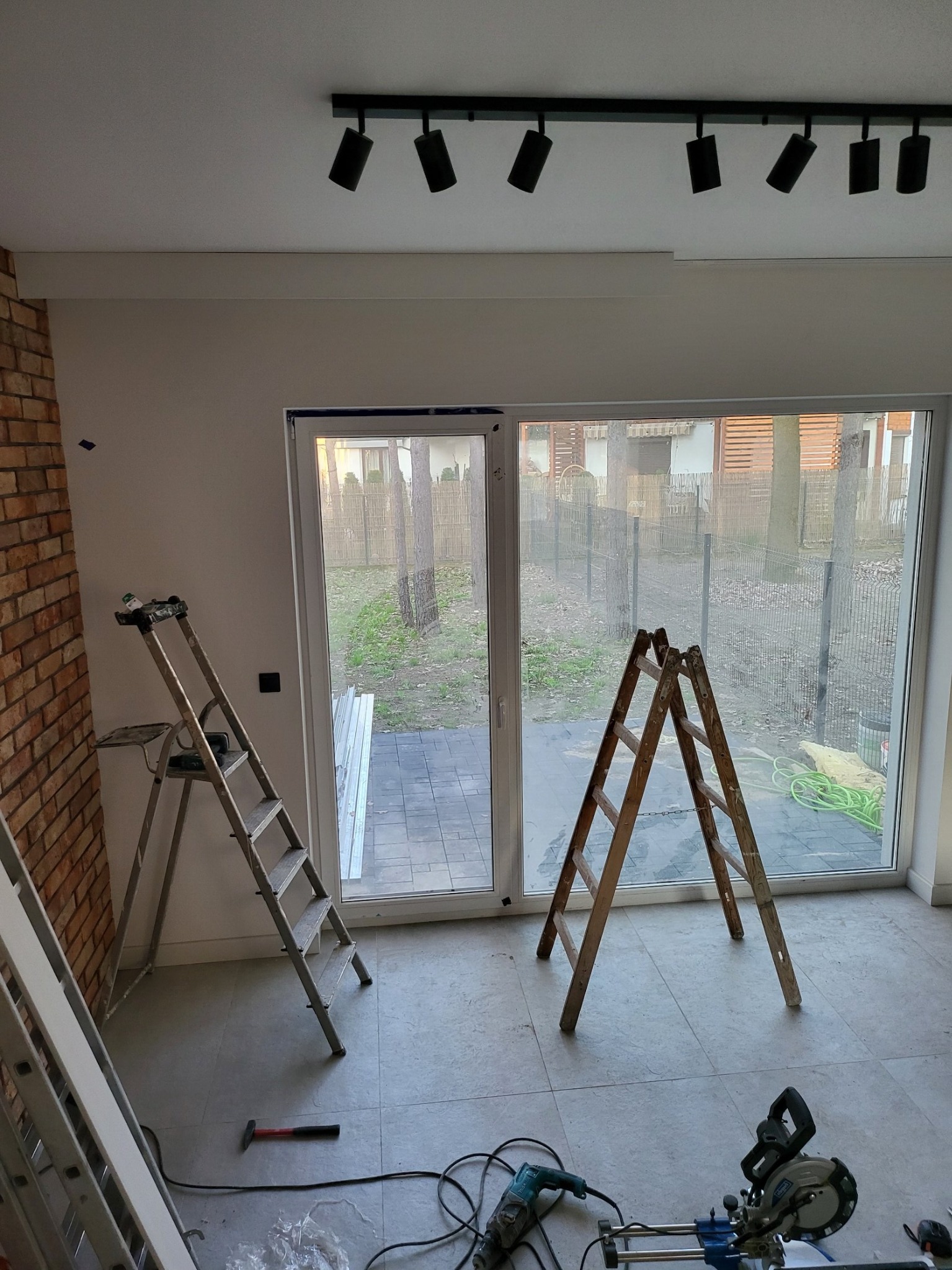 Wnętrze w trakcie prac: widoczne dwie drabiny, narzędzia (wiertarka, piła), fragment ściany z cegły, duże okno przesuwne z widokiem na zewnątrz, oświetlenie sufitowe szynowe.