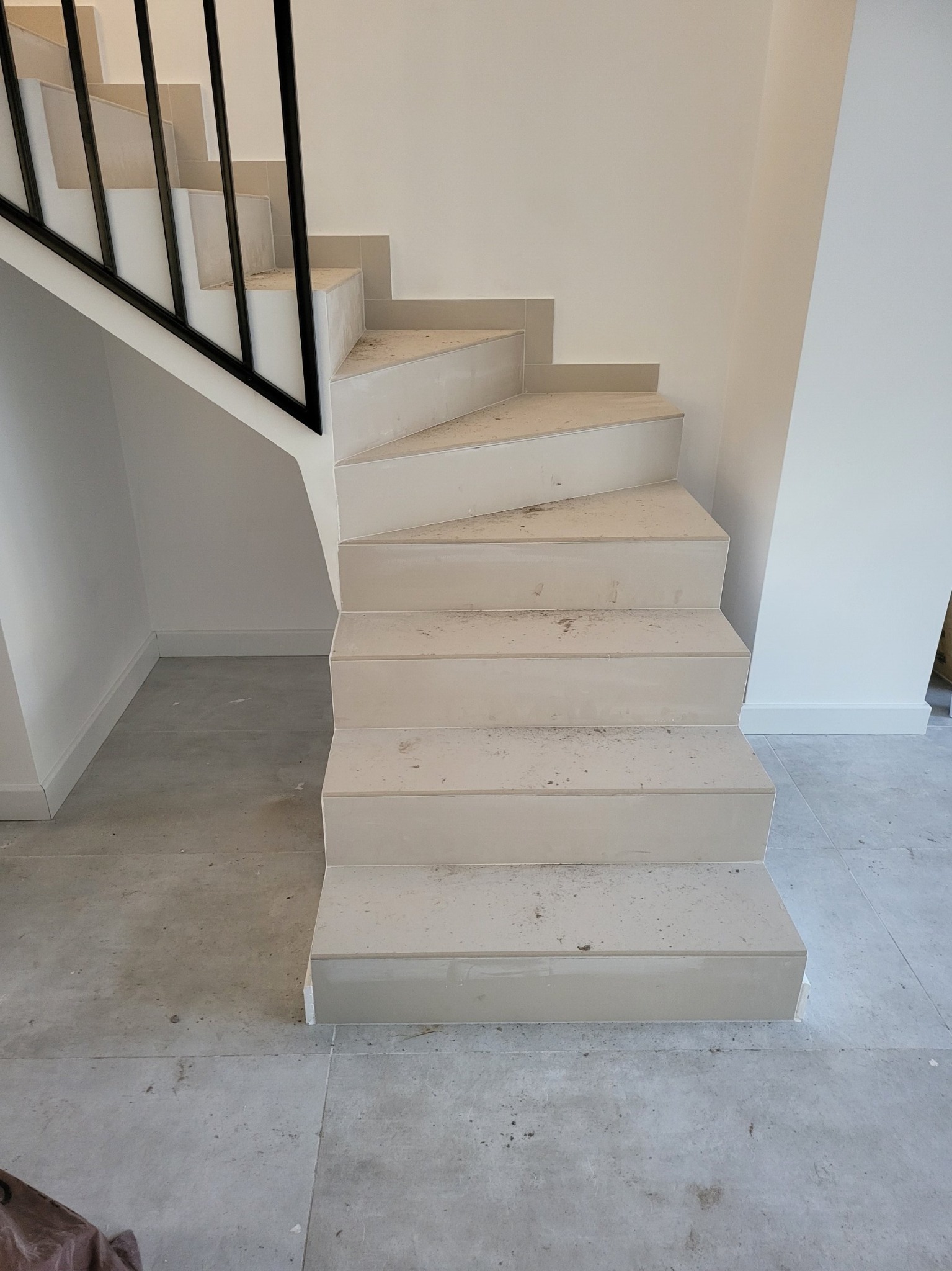 Schody wykończone jasnymi płytkami, z czarną metalową balustradą, widoczne ślady po pracach budowlanych.