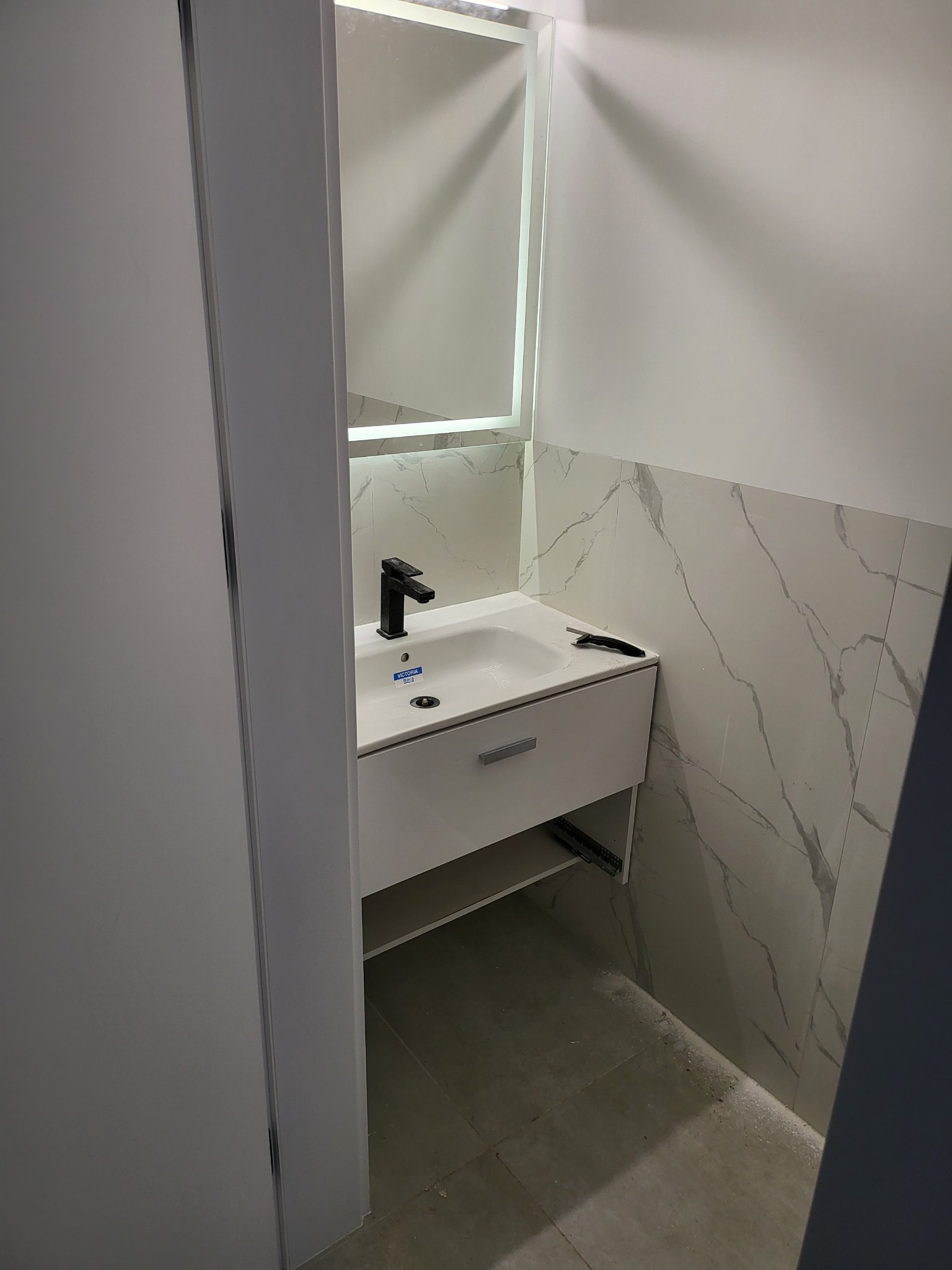 Nowoczesna łazienka z umywalką nablatową, czarną armaturą i podświetlanym lustrem, wykończona płytkami imitującymi marmur, widoczny nóż do tapet na blacie umywalki.