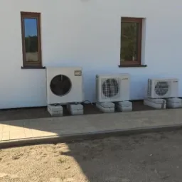 Trzy jednostki zewnętrzne pomp ciepła Mitsubishi na betonowych podstawach, zainstalowane przy białej ścianie budynku z trzema oknami o brązowych ramach i roletach.