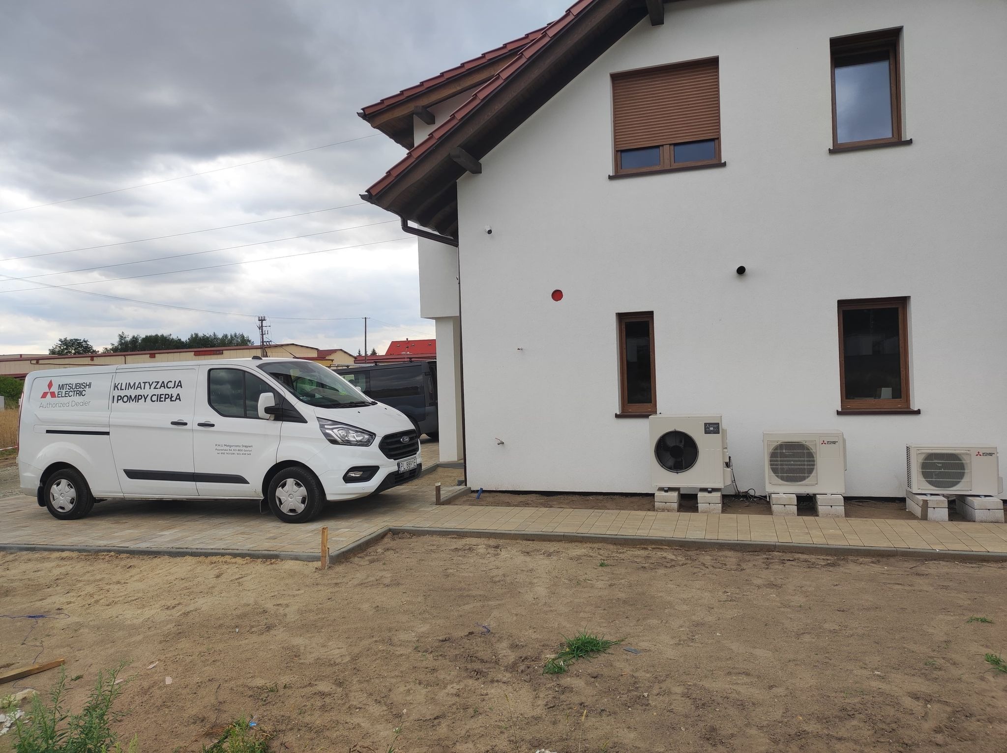 Biały van z napisem 'Klimatyzacja i Pompy Ciepła' zaparkowany obok białego domu z trzema zewnętrznymi jednostkami klimatyzacji na betonowych blokach, widok z poziomu gruntu na tle pochmurnego nieba.