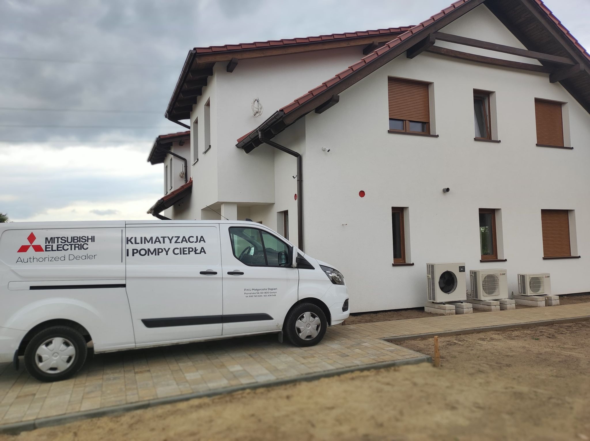 Biały budynek z trzema zewnętrznymi jednostkami klimatyzacyjnymi i zaparkowanym białym vanem z logo Mitsubishi Electric oraz napisem 'Klimatyzacja i Pompy Ciepła'.