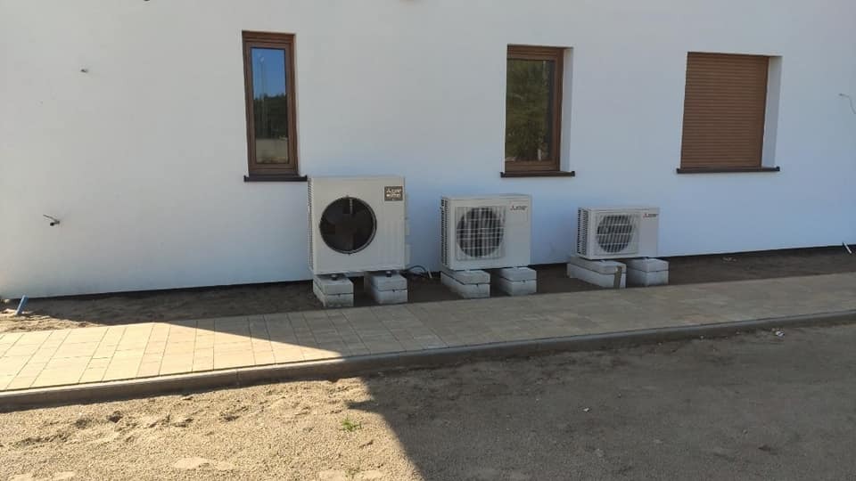 Trzy jednostki zewnętrzne pomp ciepła Mitsubishi na betonowych podstawach, zainstalowane przy białej ścianie budynku z trzema oknami o brązowych ramach i roletach.