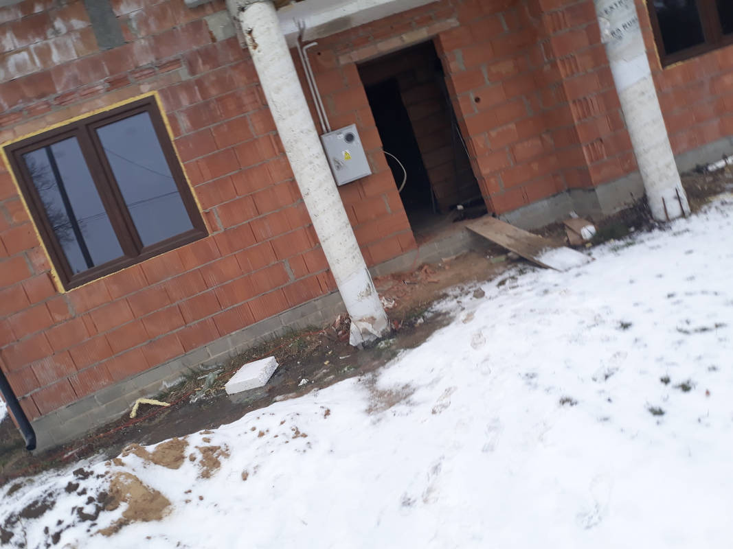 Surowy stan budynku z czerwonej cegły w Wierbce, z widoczną skrzynką elektryczną na zewnątrz i oknem z brązową ramą, częściowo zasypany śniegiem.