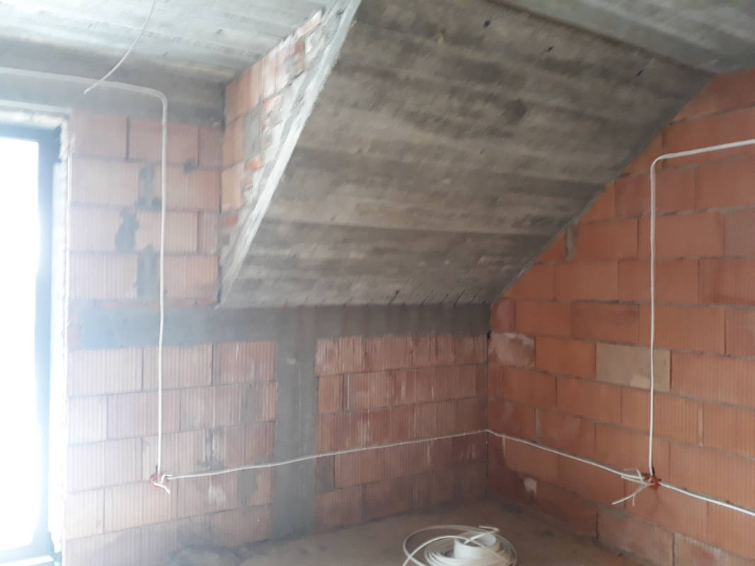 Surowy stan poddasza z widoczną instalacją elektryczną: przewody poprowadzone po ceglanych ścianach pod betonowym stropem, zwój kabla na podłodze.