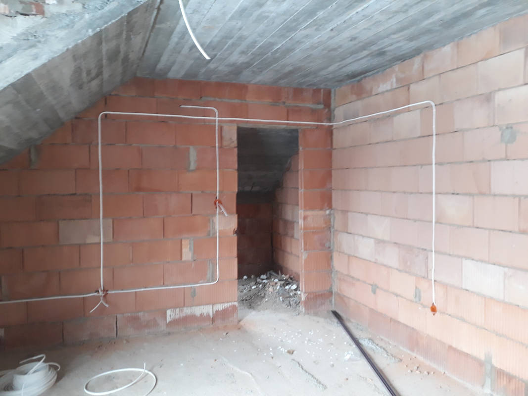 Surowy stan poddasza z widocznymi przewodami elektrycznymi poprowadzonymi po ścianach z czerwonej cegły dziurawki, z betonowym stropem i otworem drzwiowym prowadzącym do ciemnego pomieszczenia.