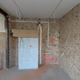 Wnętrze w trakcie remontu z widocznymi przewodami elektrycznymi prowadzonymi po ścianie z cegły, zabezpieczona skrzynka elektryczna i surowy stan wykończenia.