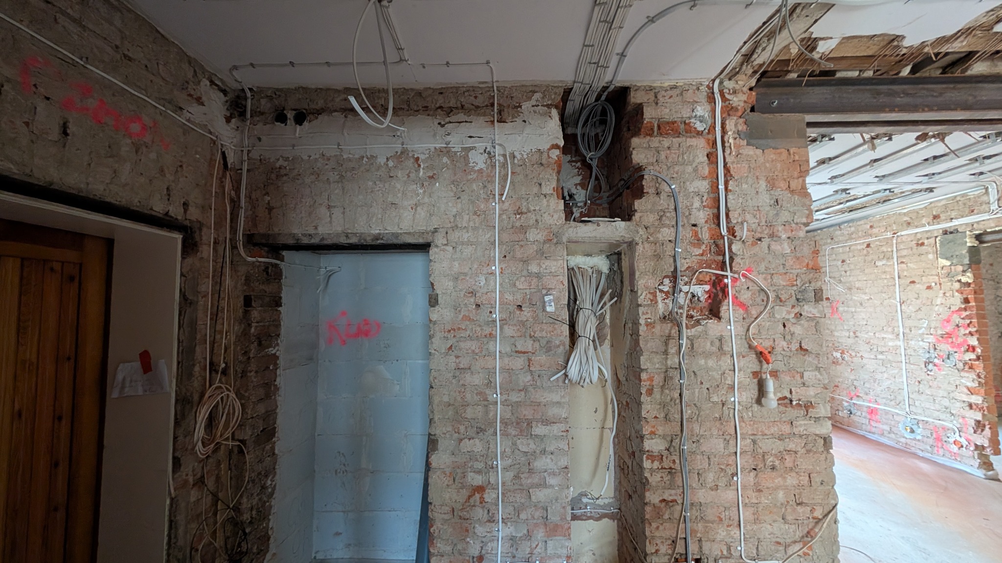 Wnętrze w trakcie remontu z odsłoniętą cegłą, widoczne przewody elektryczne prowadzone po ścianach i suficie, otwór w ścianie z wiązką przewodów zabezpieczoną sznurkiem, czerwone oznaczenia...
