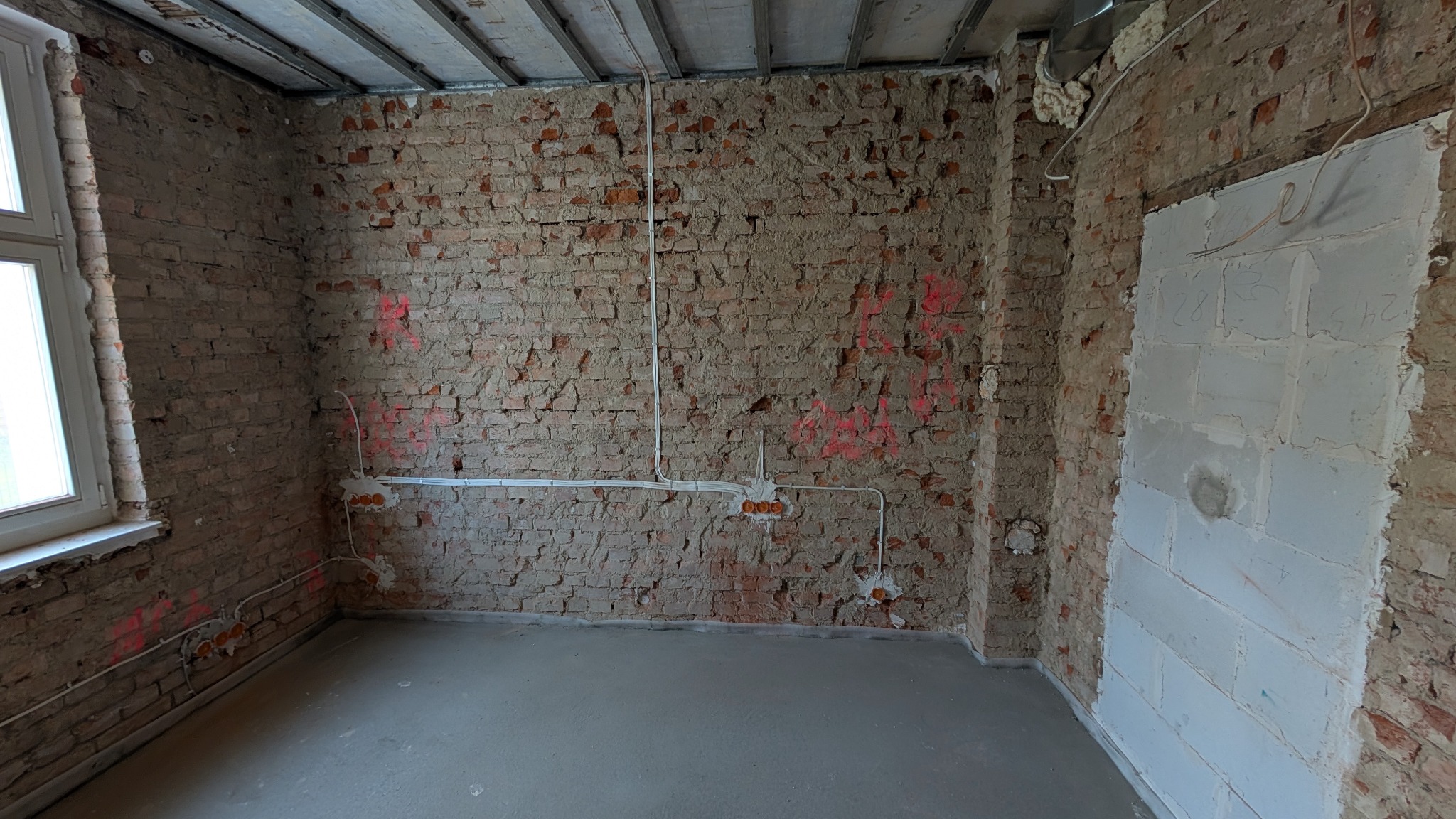 Surowy stan wnętrza w trakcie remontu: widoczne ceglane ściany z fragmentami tynku, nowa instalacja elektryczna natynkowa z puszkami, częściowo zamurowane otwory, betonowa wylewka na podłodze...