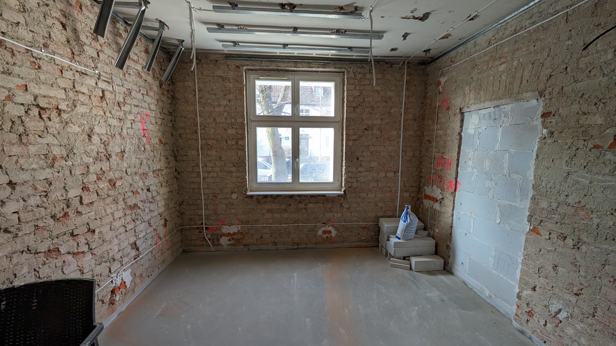 Surowy pokój z odsłoniętą cegłą, widoczne przewody elektryczne, metalowe profile pod sufitem, okno z widokiem na drzewo, worki z zaprawą na podłodze, przygotowany otwór drzwiowy.