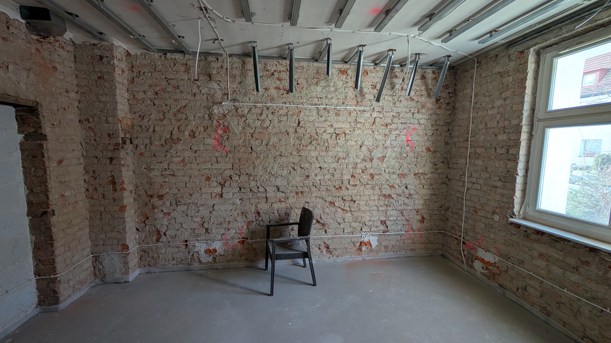 Surowe wnętrze w trakcie remontu z odsłoniętą cegłą, widocznymi przewodami elektrycznymi pod sufitem i na ścianach, oraz metalowymi elementami konstrukcyjnymi sufitu podwieszanego. Na środku stoi...