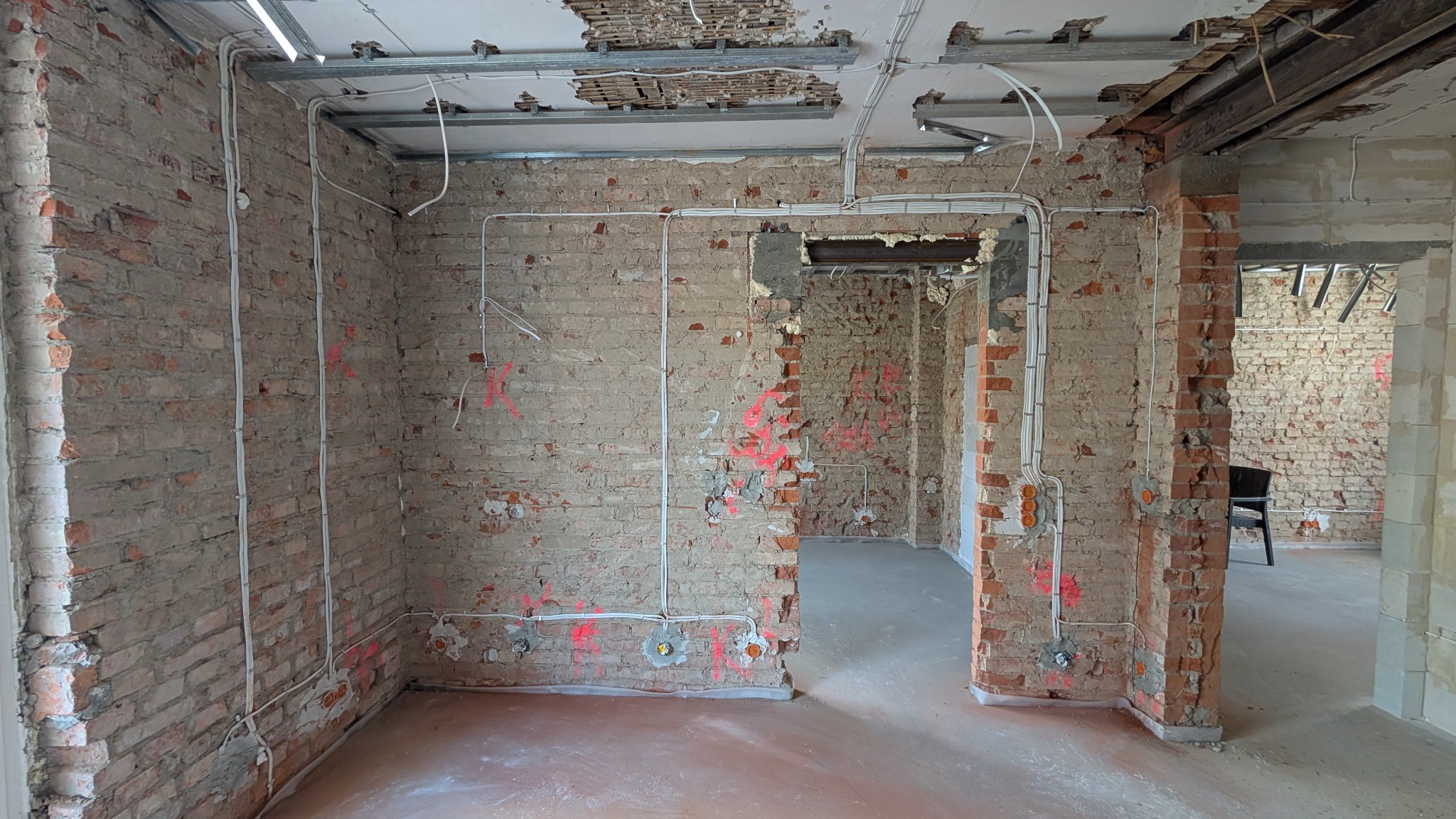 Wnętrze w trakcie remontu: odsłonięta cegła, nowe przewody elektryczne prowadzone po ścianach, oznaczenia instalacji czerwoną farbą, odsłonięta konstrukcja sufitu.