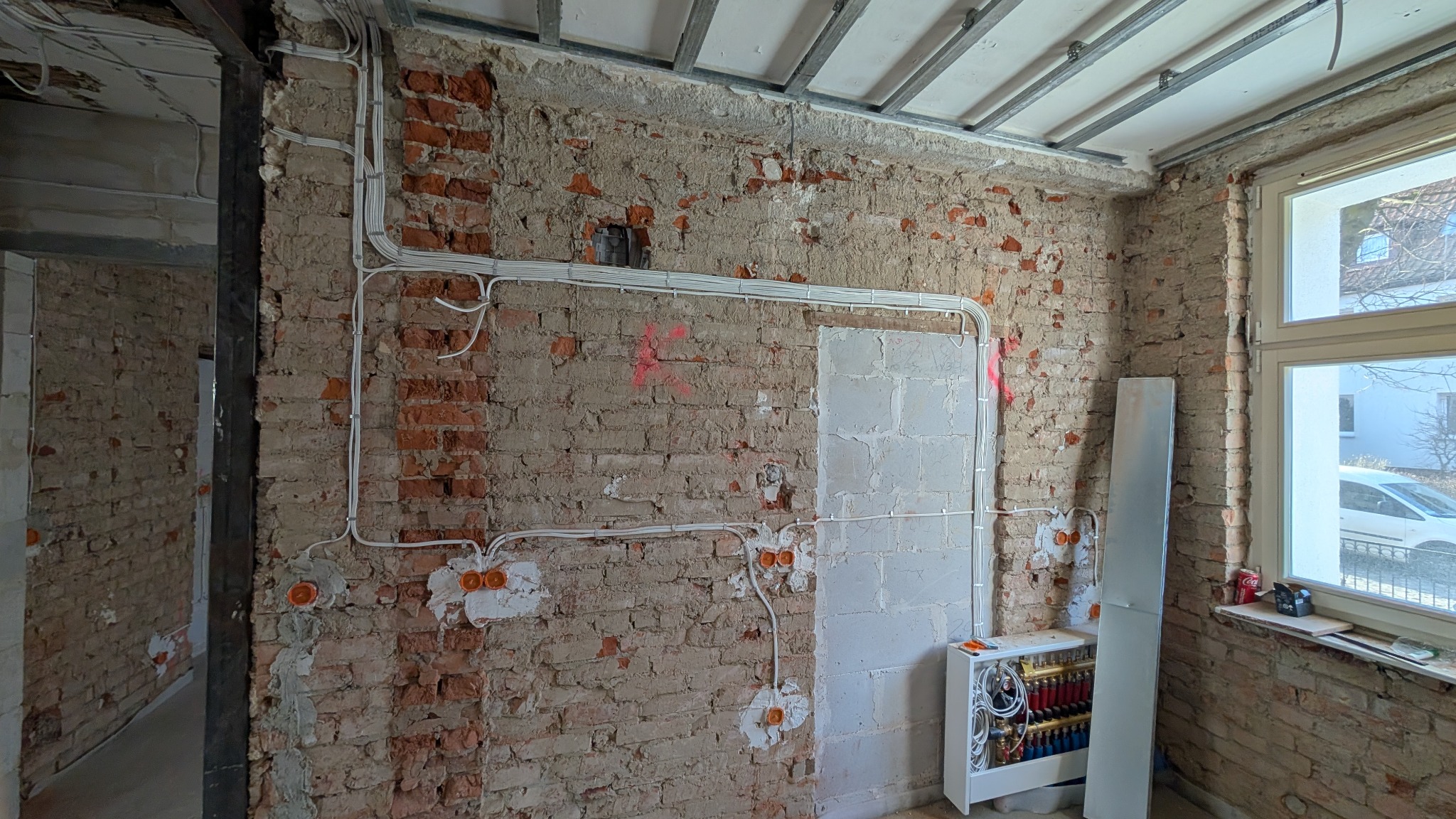 Surowa ściana z cegły z widocznymi przewodami elektrycznymi, puszkami instalacyjnymi i instalacją ogrzewania podłogowego w trakcie remontu.