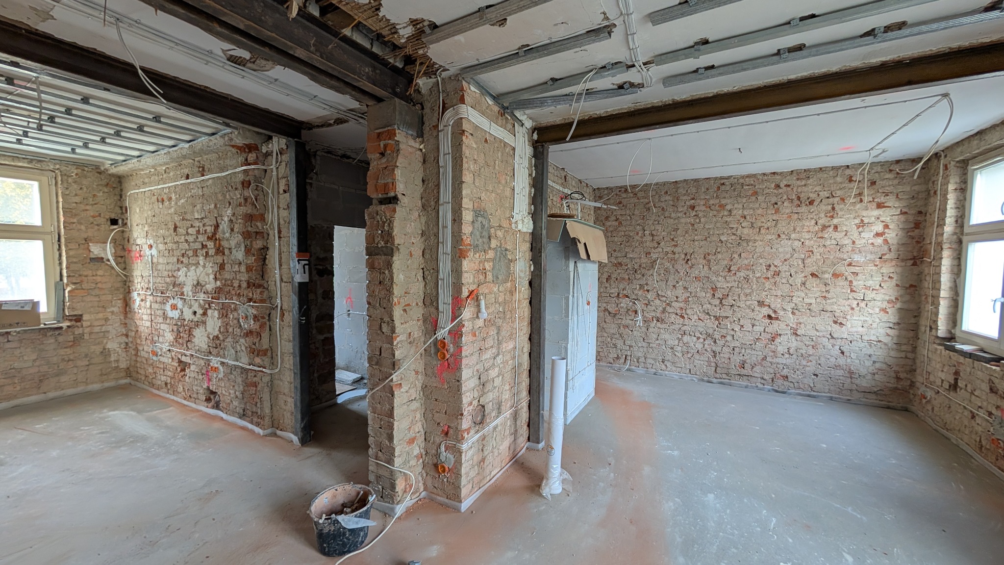 Wnętrze w trakcie remontu z odsłoniętą cegłą, widoczne kable elektryczne w peszlach prowadzone po ścianach i pod sufitem, metalowe profile podwieszanego sufitu, surowe ściany i podłoga, kubeł...