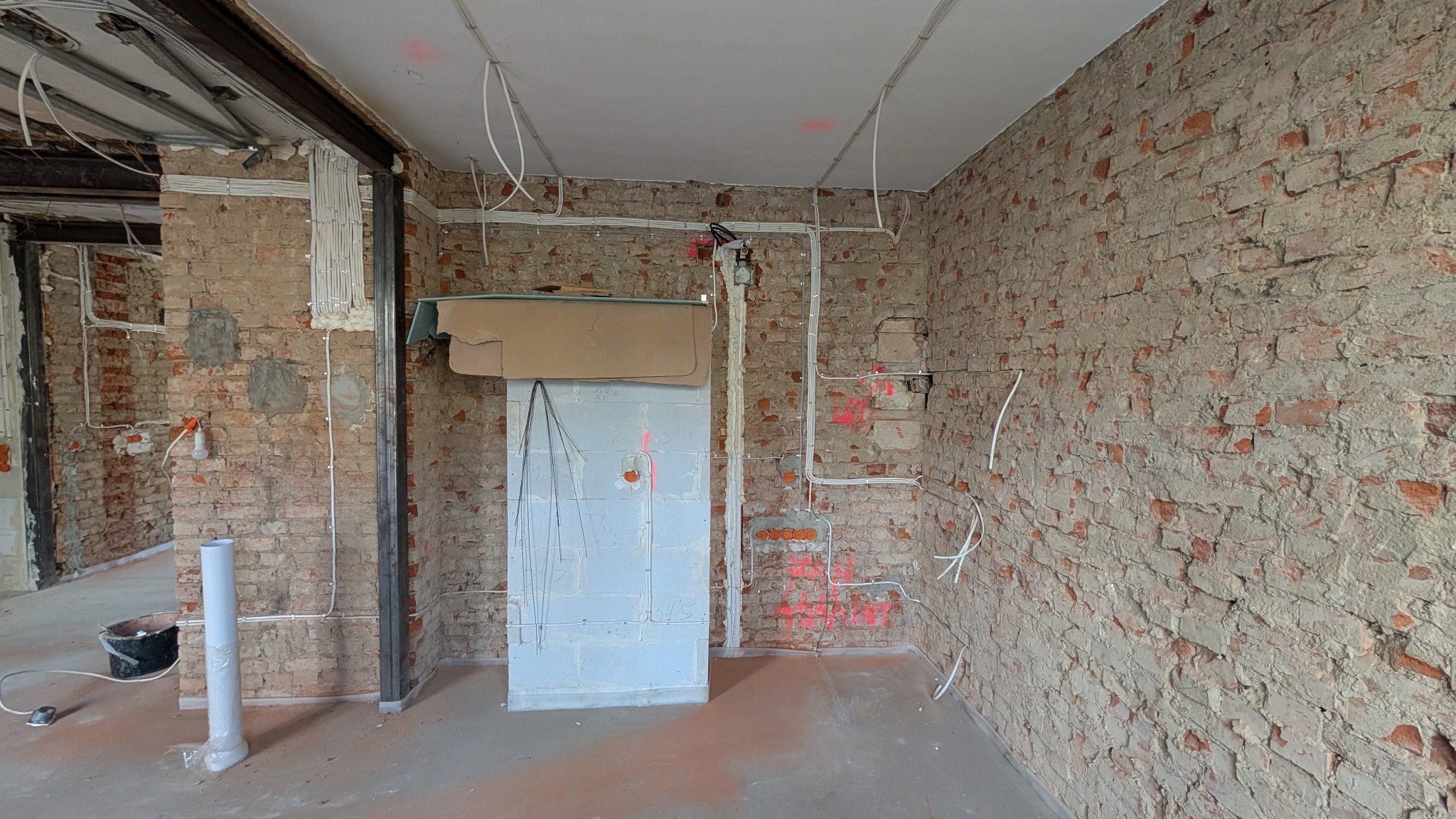 Wnętrze w trakcie remontu z widocznymi przewodami elektrycznymi prowadzonymi po ścianie z cegły, zabezpieczona skrzynka elektryczna i surowy stan wykończenia.