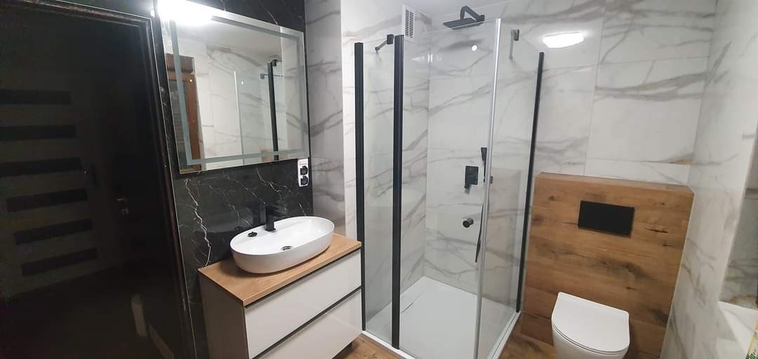 Nowoczesna łazienka po remoncie w Kędzierzynie-Koźlu: kabina prysznicowa z czarnymi profilami, umywalka nablatowa na szafce z drewnianym blatem, lustro z podświetleniem LED i marmurowe płytki.