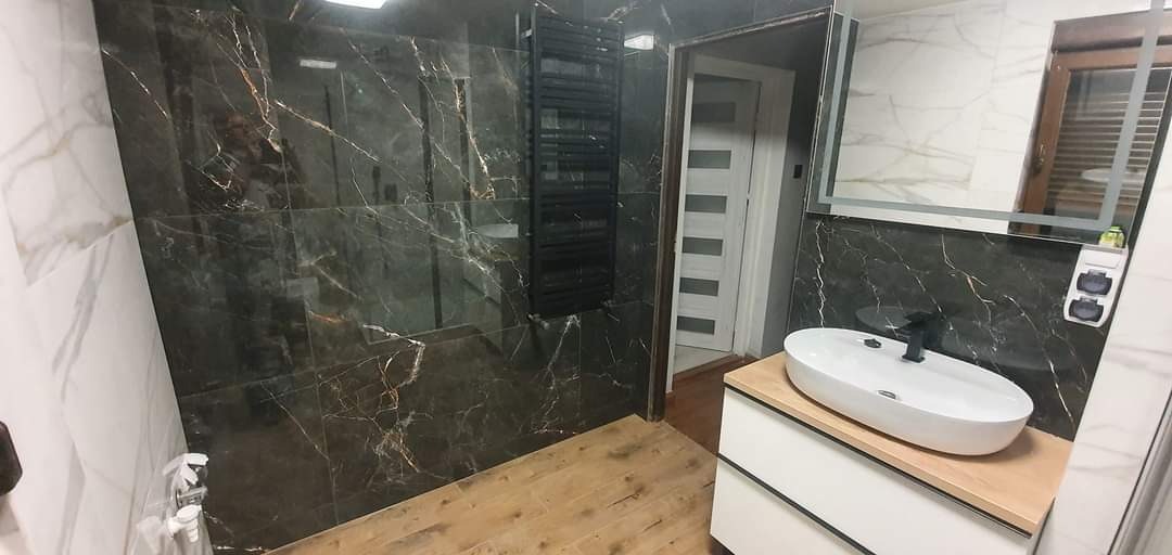 Wykończona łazienka z czarnymi płytkami imitującymi marmur z złotymi żyłami, drewnianą podłogą, białym blatem umywalki i grzejnikiem drabinkowym.