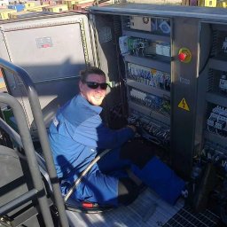Lema electro - Uśmiechnięty elektryk w kombinezonie roboczym i okularach przeciwsłonecznych, siedzący wewnątrz szafy sterowniczej z licznymi przewodami i elementami elektrycznymi, na tle kontenerów portowych.