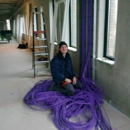 Lema electro - Uśmiechnięty elektryk siedzi na stercie fioletowych kabli w surowym wnętrzu budynku w trakcie instalacji elektrycznej, drabina oparta o ścianę w tle.