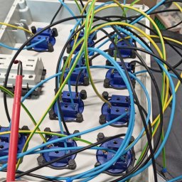 Lema electro - Rozdzielnica elektryczna w trakcie montażu: widoczne niebieskie gniazda, przewody w izolacji żółto-zielonej, niebieskiej i czarnej, oraz czerwony próbnik napięcia w dłoni montera.