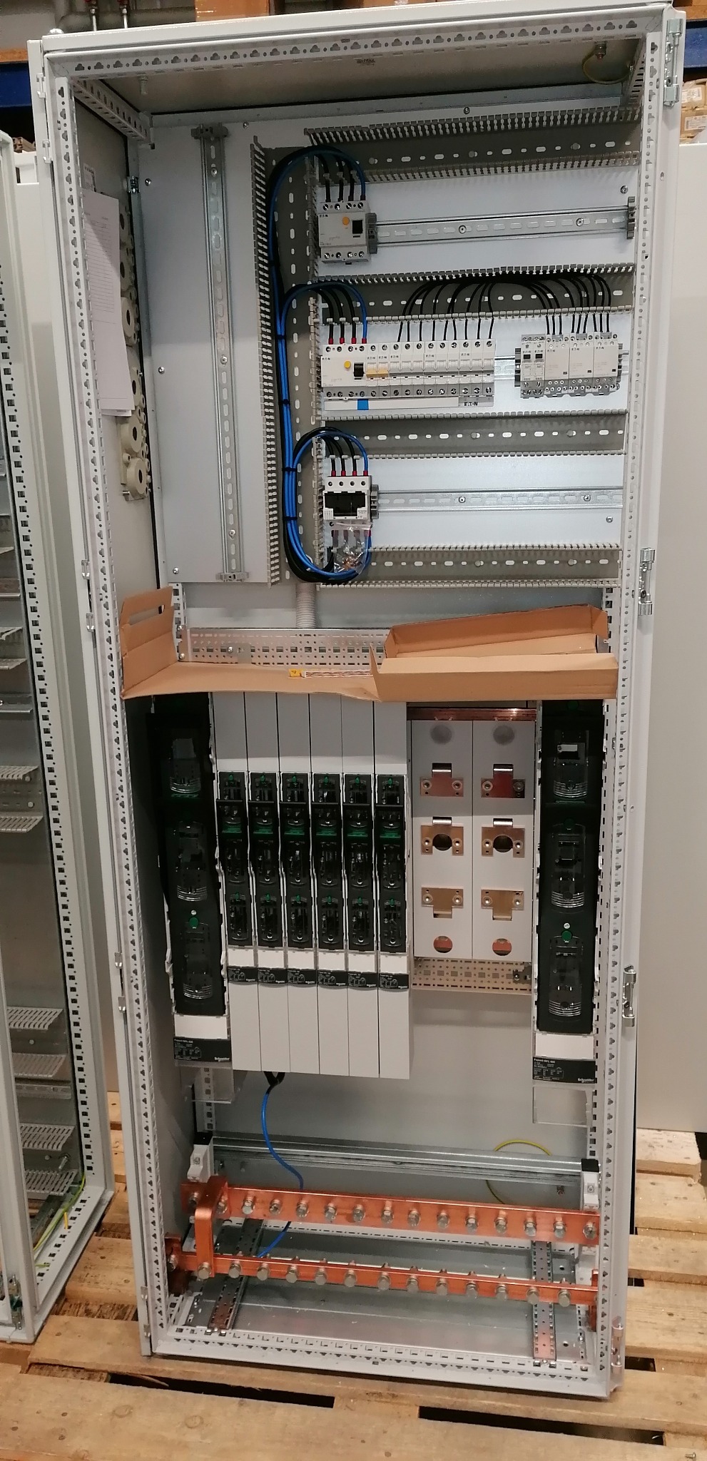 Otwarta szafa elektryczna w trakcie montażu, widoczne zabezpieczenia, przewody, szyny montażowe i elementy sterujące, częściowo zabezpieczone kartonem.