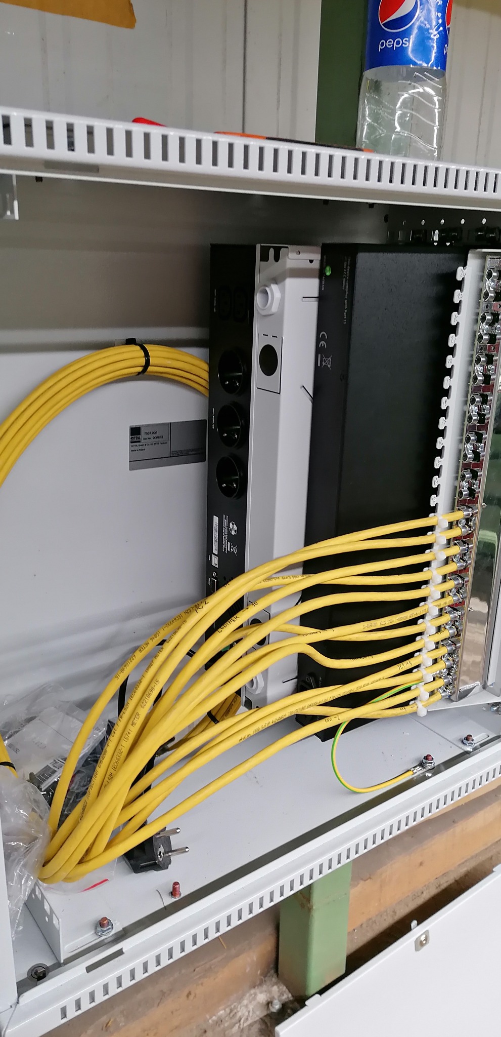 Wnętrze szafy elektrycznej z ułożonymi żółtymi kablami, listwą z gniazdami, modułem z zaciskami śrubowymi, oraz butelką Pepsi na górze.