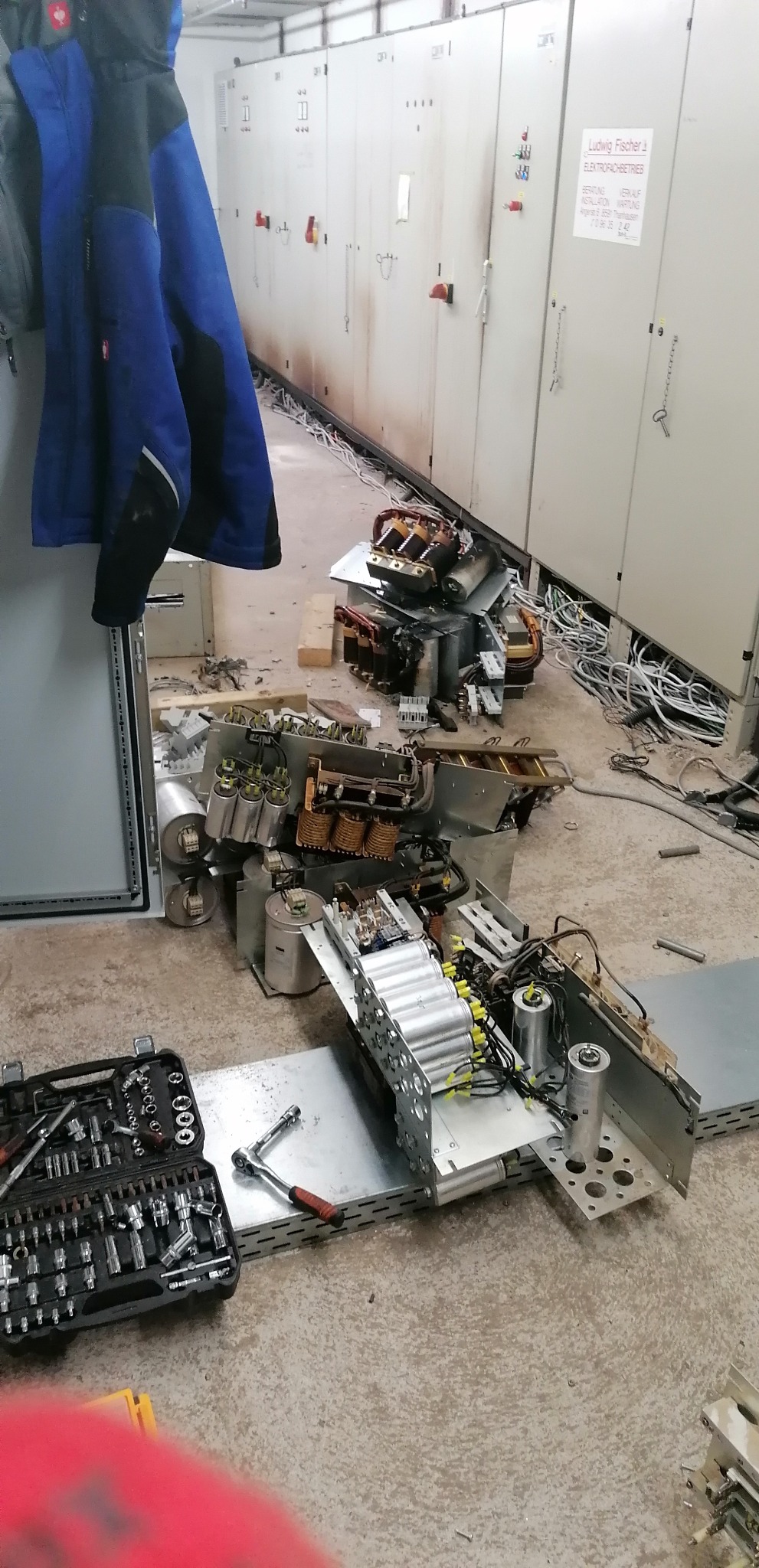 Demontaż uszkodzonej rozdzielnicy elektrycznej w Chrzanowie. Na podłodze rozłożone części, widoczny otwarty kufer z narzędziami i elementy instalacji. Tło stanowią szafy sterownicze.