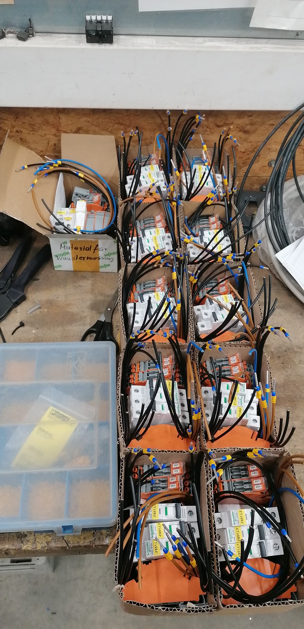 Przygotowane moduły elektryczne w kartonowych pudełkach, okablowane i gotowe do montażu, z widocznymi oznaczeniami Schneider Electric, obok skrzynka z elementami montażowymi i narzędzia.