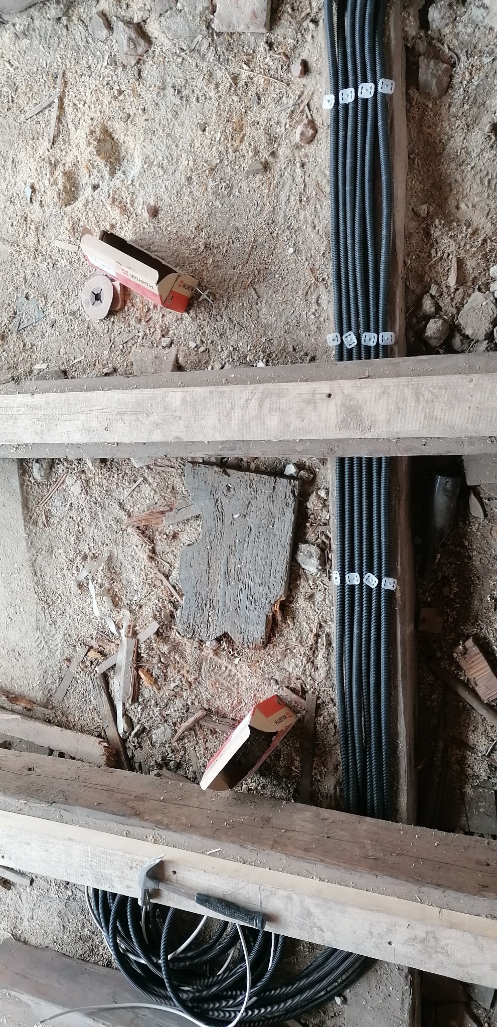 Wiązki czarnych przewodów elektrycznych ułożone w bruzdach w podłodze, przytwierdzone białymi uchwytami, obok leżą otwarte pudełka po kołkach marki Wurth i młotek na drewnianej belce, widoczne...