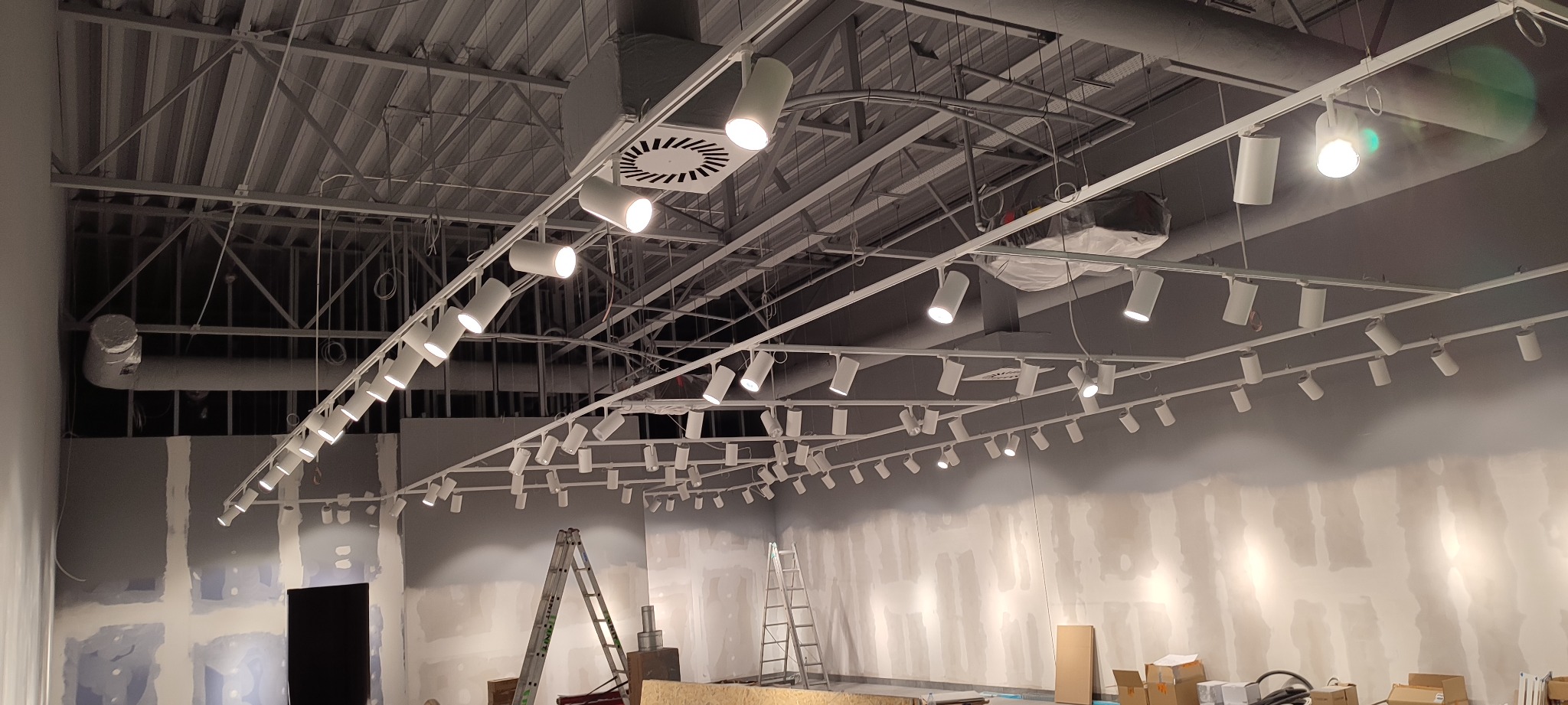 Montaż szynoprzewodów z reflektorami w surowym wnętrzu, widoczne drabiny i materiały budowlane.