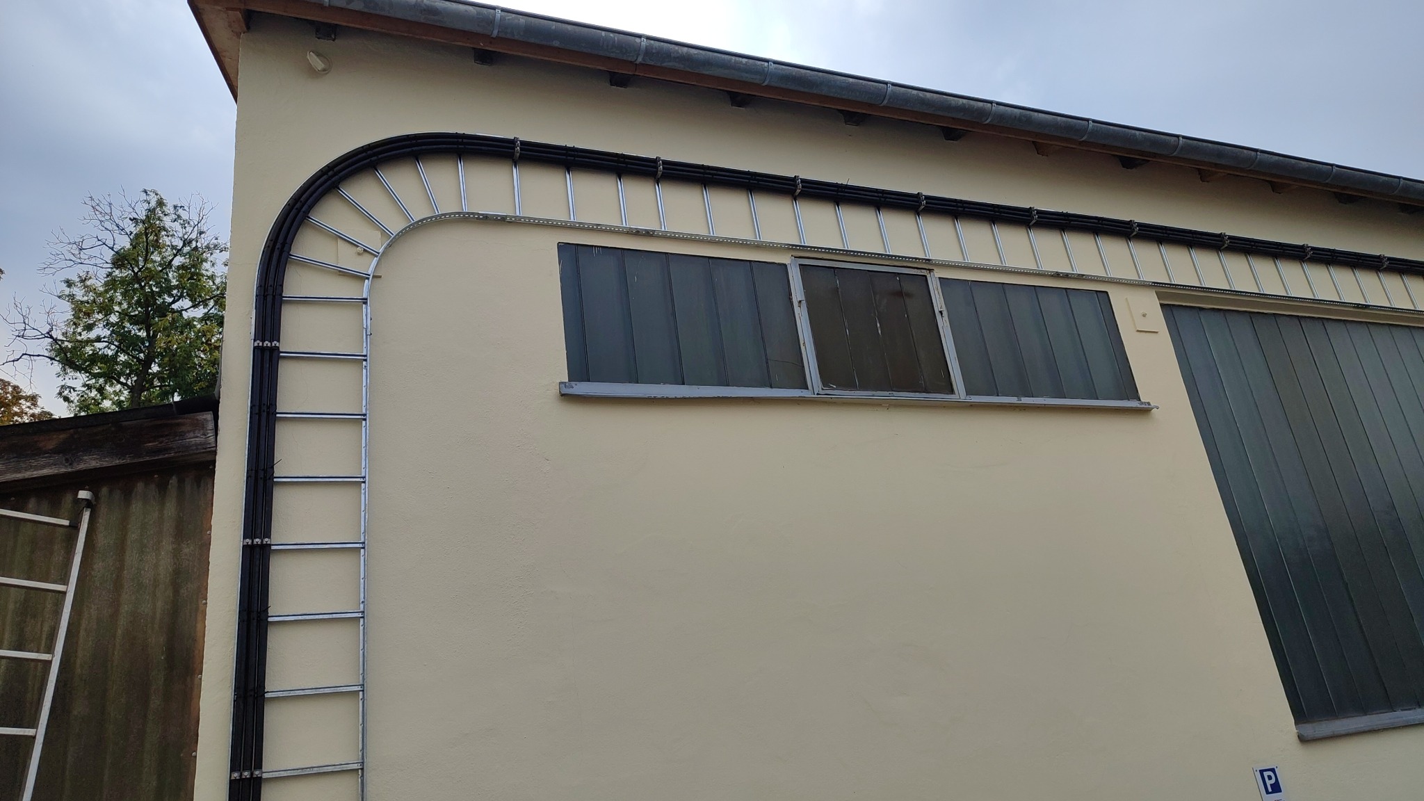 Zewnętrzna instalacja kabli elektrycznych w korytach i rurach na elewacji budynku, z widoczną drabinką i oknami.