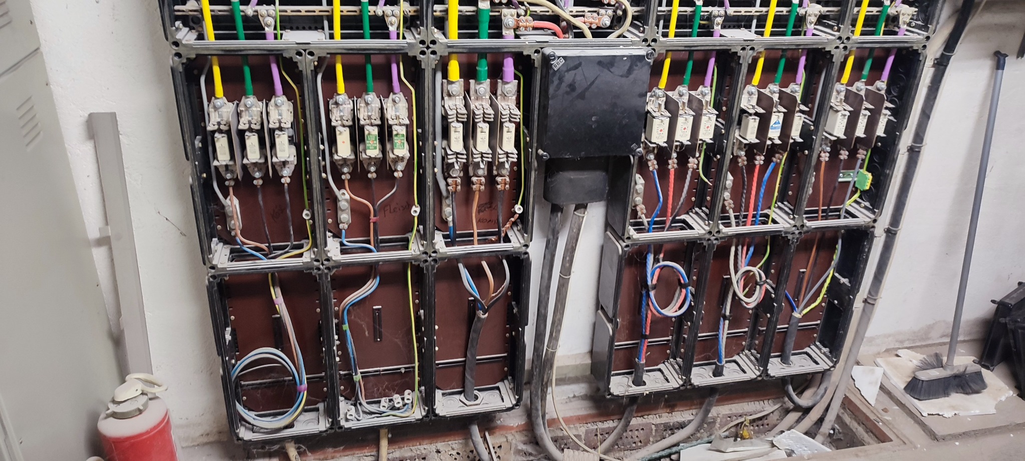 Rozbudowana skrzynka elektryczna z bezpiecznikami i kolorowymi przewodami, umieszczona w piwnicy, widoczny sprzęt gaśniczy i otwarta studzienka rewizyjna.