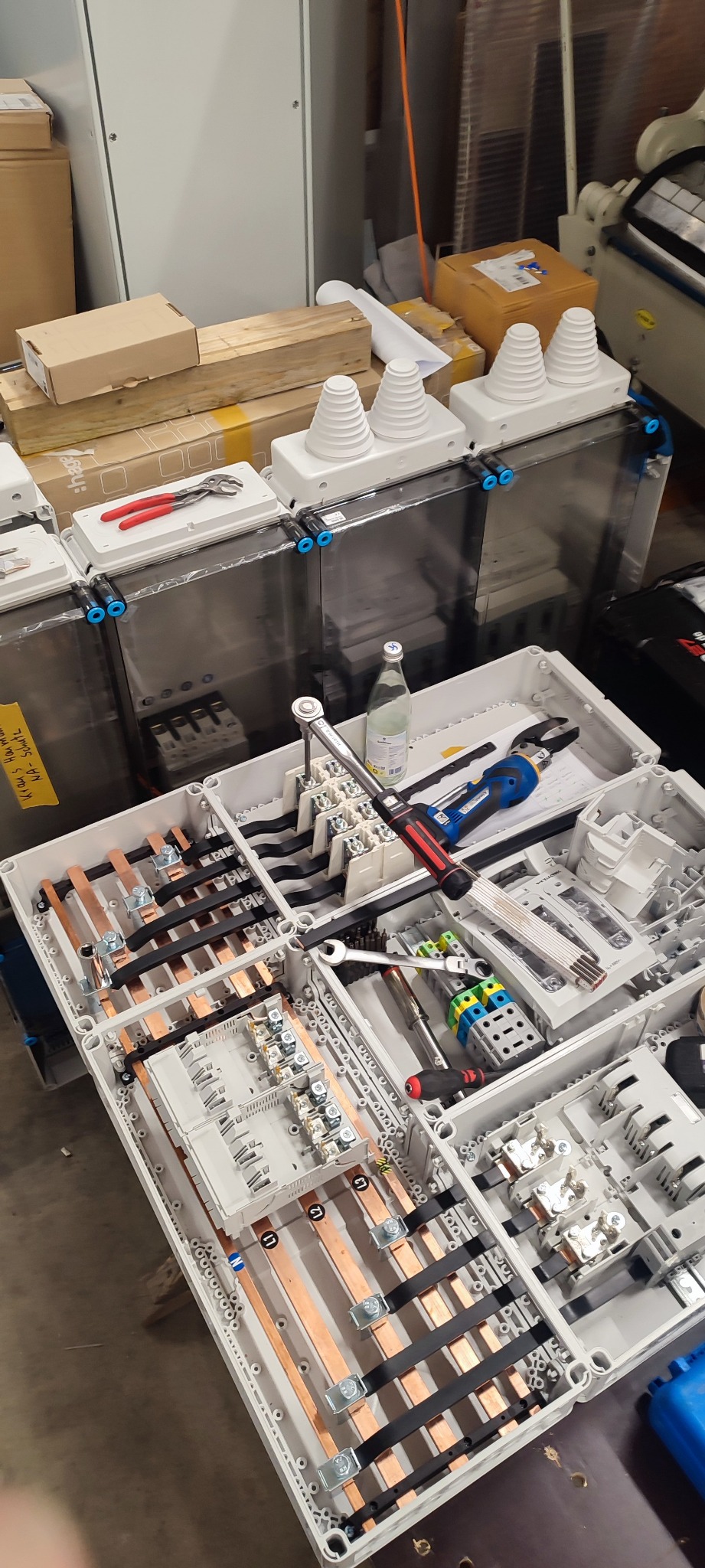 Rozdzielnica elektryczna w trakcie montażu, widoczne szyny miedziane, przewody, narzędzia, butelka wody i skrzynki z elementami w tle.