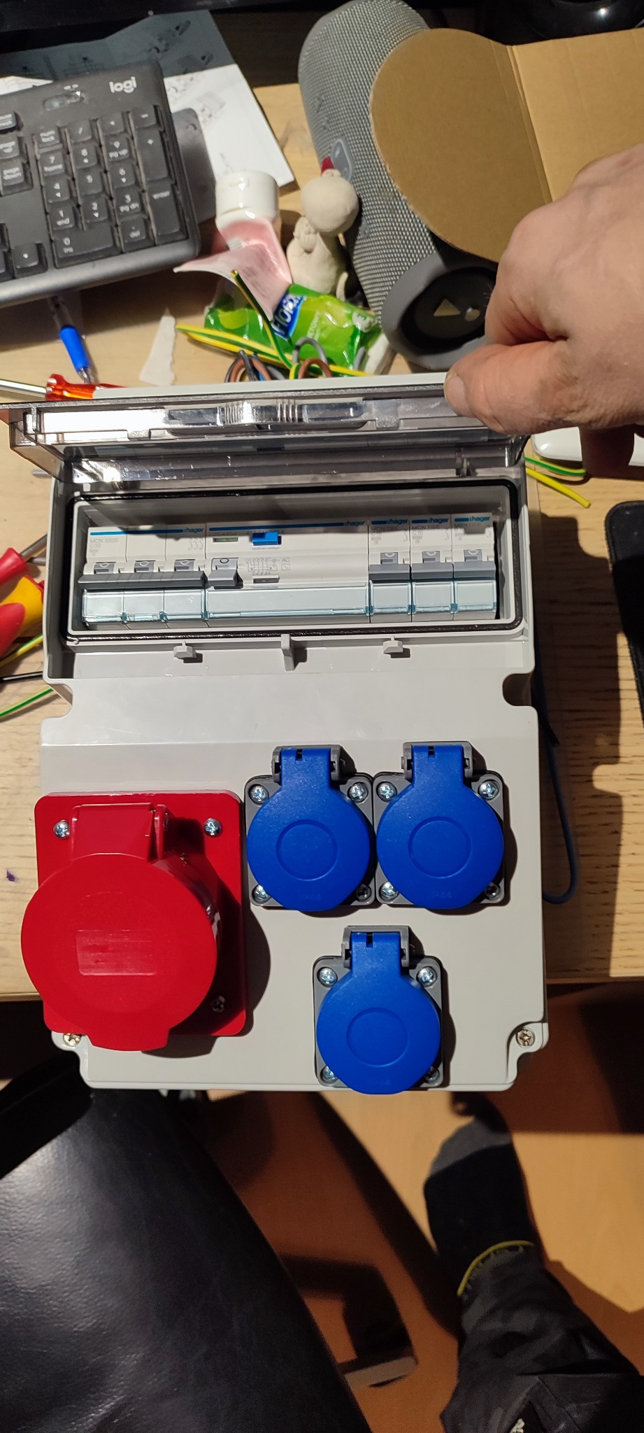 Rozdzielnica elektryczna z otwartą klapką, widoczne zabezpieczenia i gniazda siłowe w kolorze czerwonym i niebieskim, montowana na drewnianej powierzchni, w tle klawiatura i głośnik.