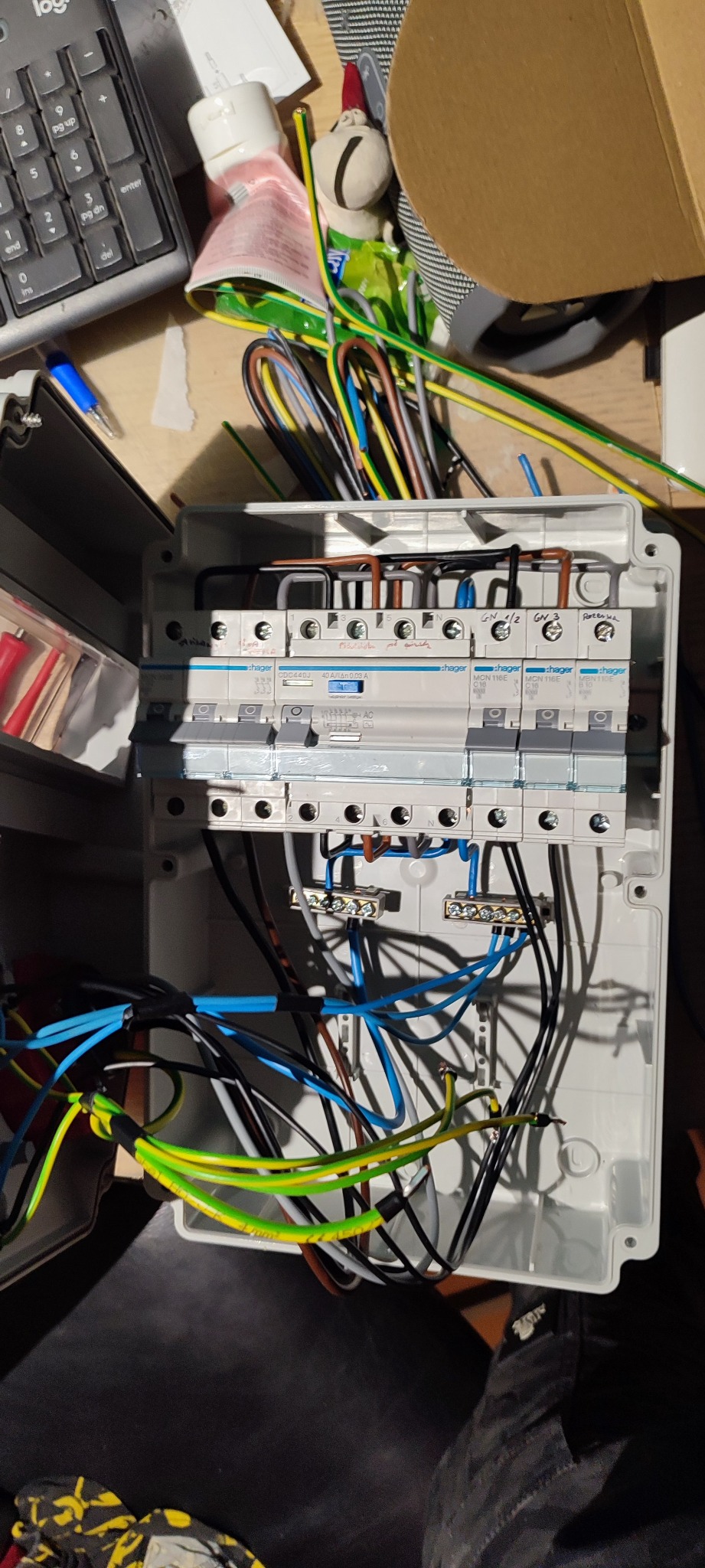 Otwarta skrzynka rozdzielcza Hager z podłączonymi przewodami o różnych kolorach, widoczne bezpieczniki i listwy zaciskowe, obok klawiatura komputera i narzędzia.