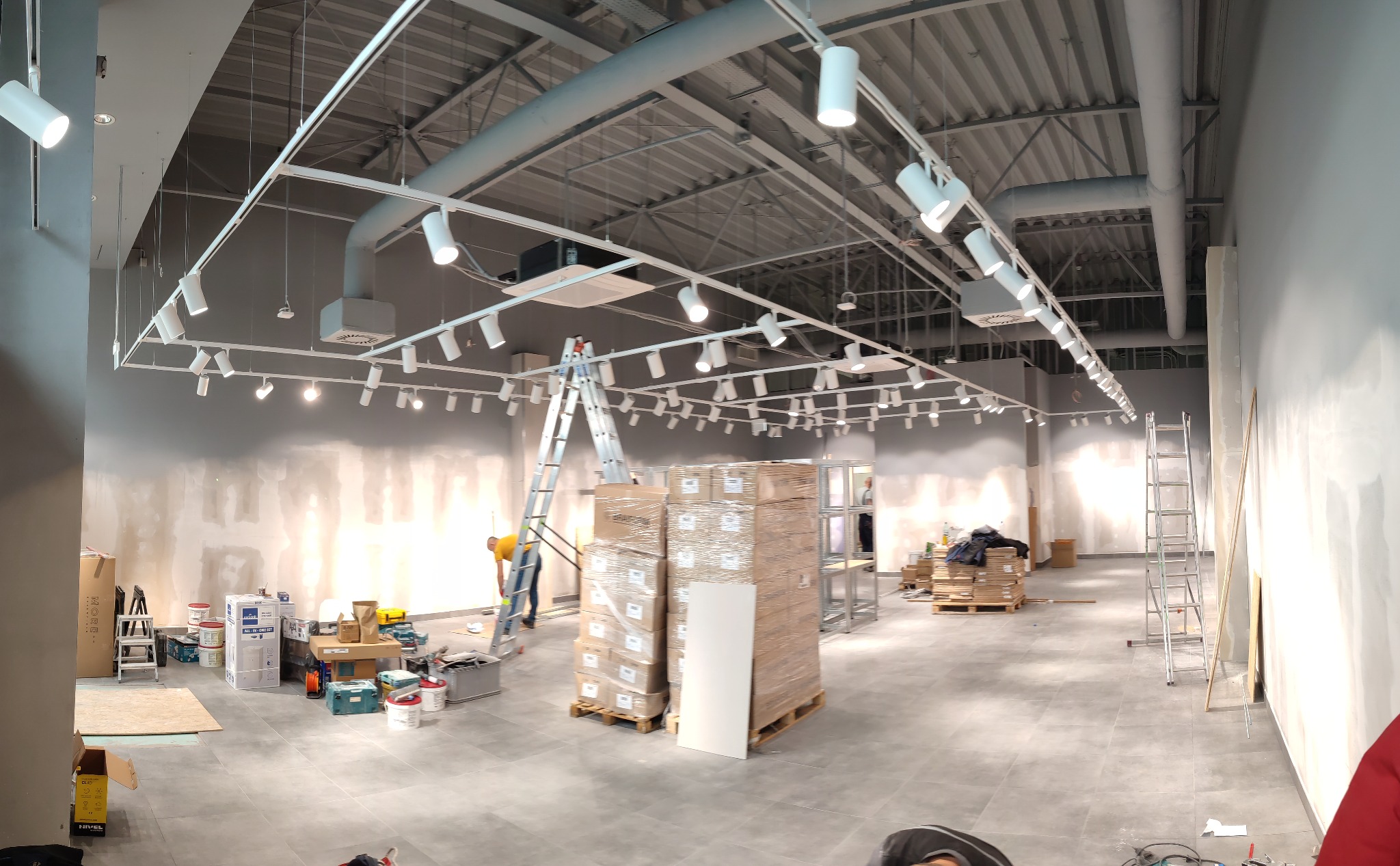 Wnętrze sklepu w trakcie montażu oświetlenia: widoczne pakiety lamp na paletach, drabiny, narzędzia i elektryk na drabinie podłączający lampy na szynoprzewodach pod sufitem.