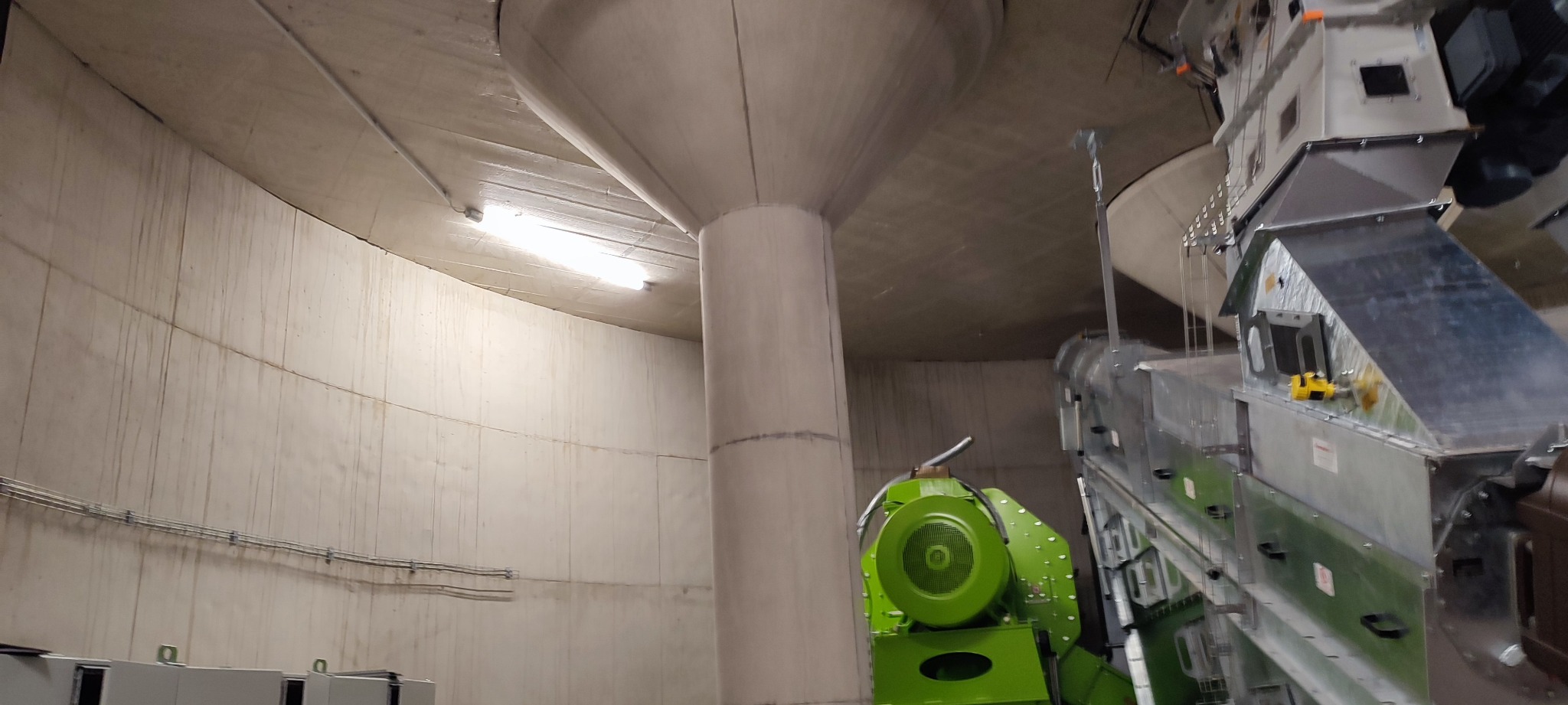 Wnętrze betonowej konstrukcji z jasnym oświetleniem, widoczny zielony wentylator przemysłowy i metalowe kanały wentylacyjne.