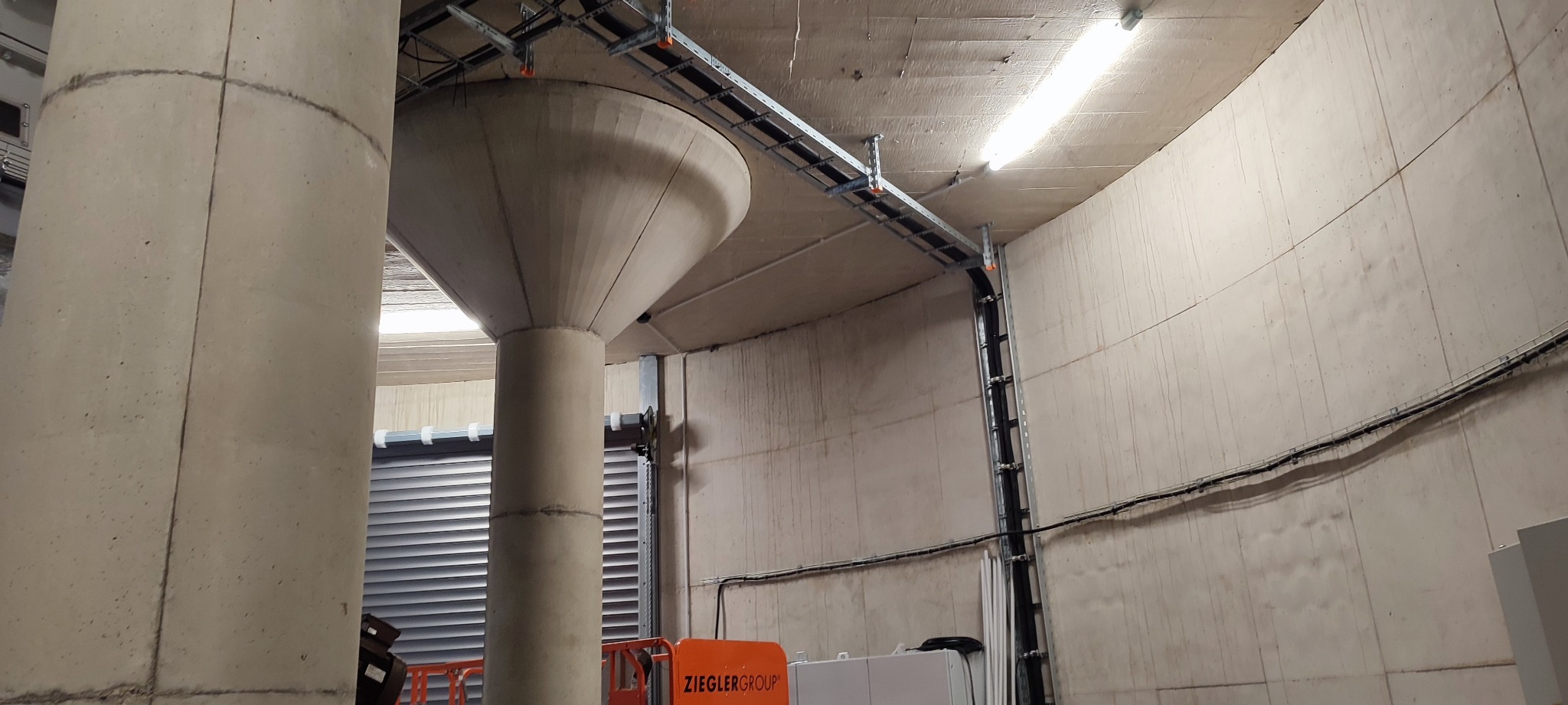 Wnętrze przemysłowe z widocznymi betonowymi kolumnami, instalacjami kablowymi pod sufitem i elementami rusztowania na pierwszym planie.