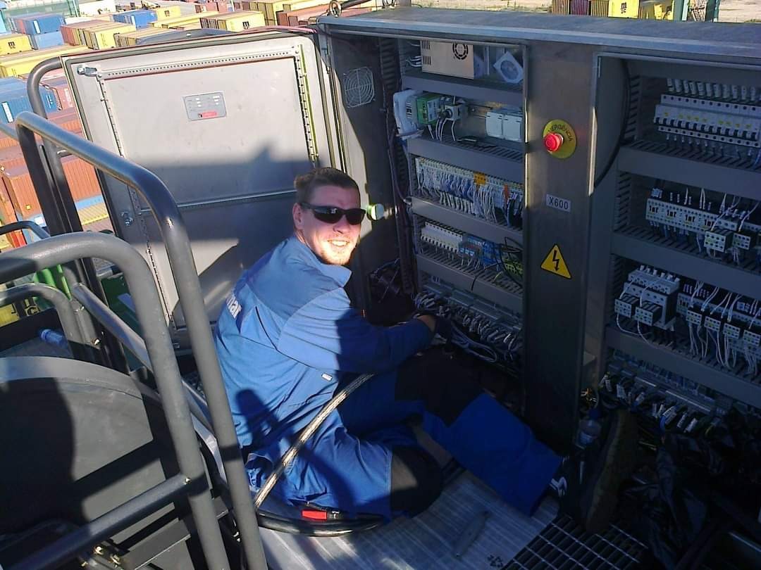 Uśmiechnięty elektryk w kombinezonie roboczym i okularach przeciwsłonecznych, siedzący wewnątrz szafy sterowniczej z licznymi przewodami i elementami elektrycznymi, na tle kontenerów portowych.